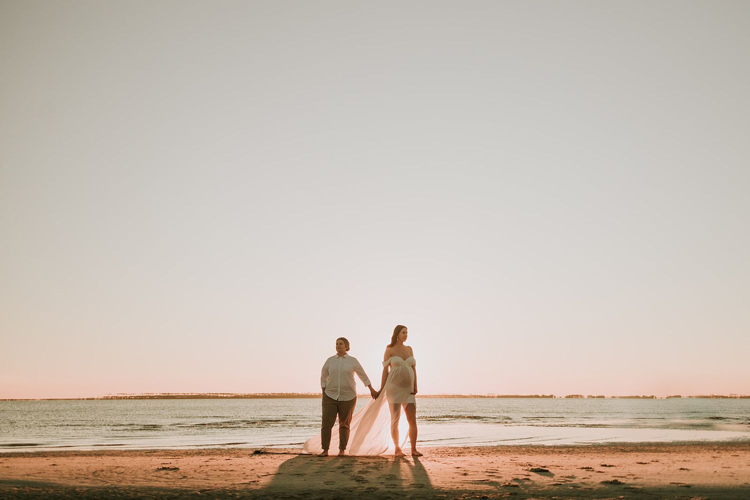 Two silhouettes stand together on a beach during sunset holding hands.