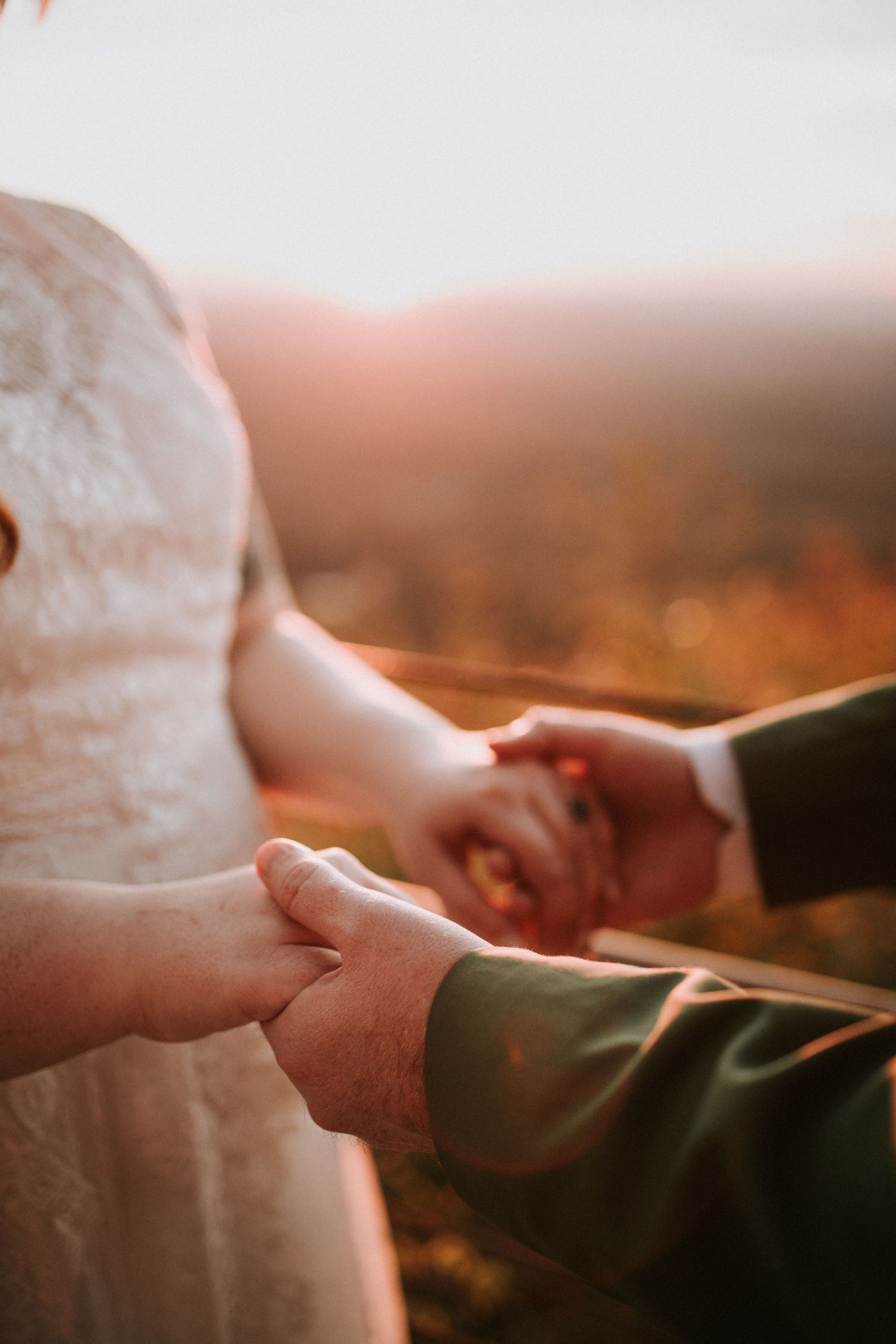 Close-up of couple holding hands during sunset with golden light and white lace dress visible in romantic moment.