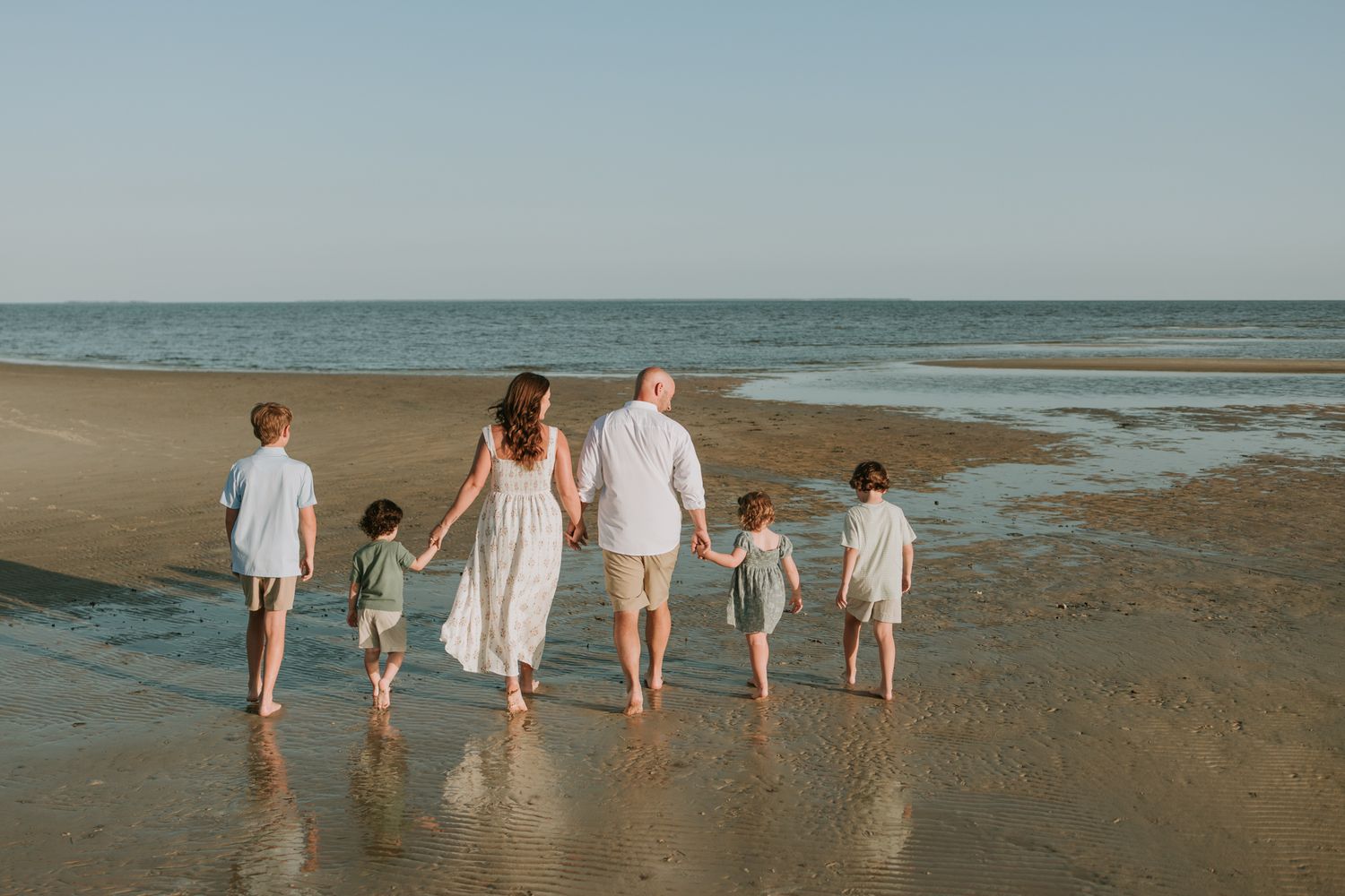 Family of six walking hand-in-hand along a sandy beach shore at sunset, dressed in matching white outfits.