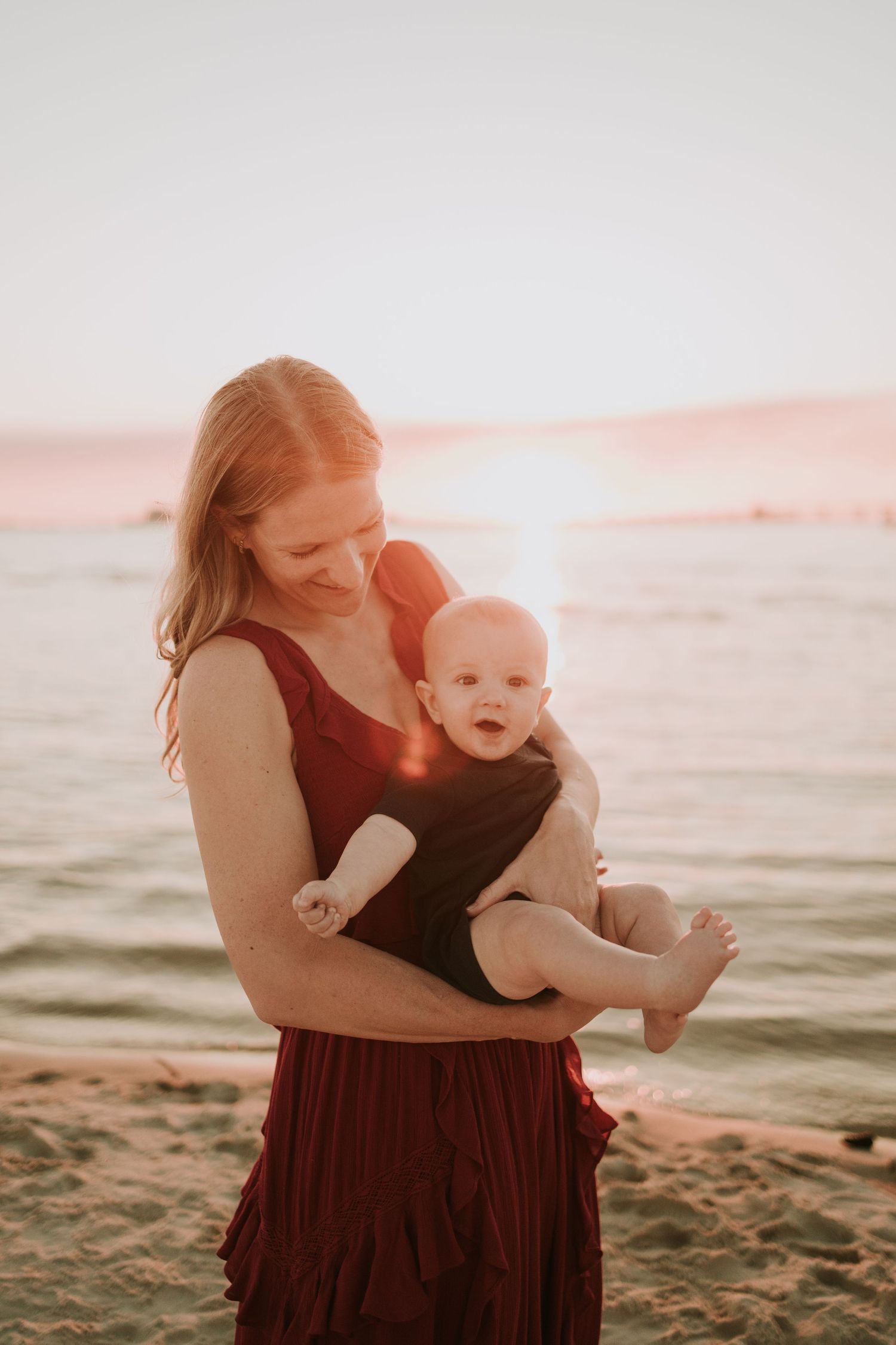 A mother in a red dress holds her baby on a sandy beach at golden hour sunset.