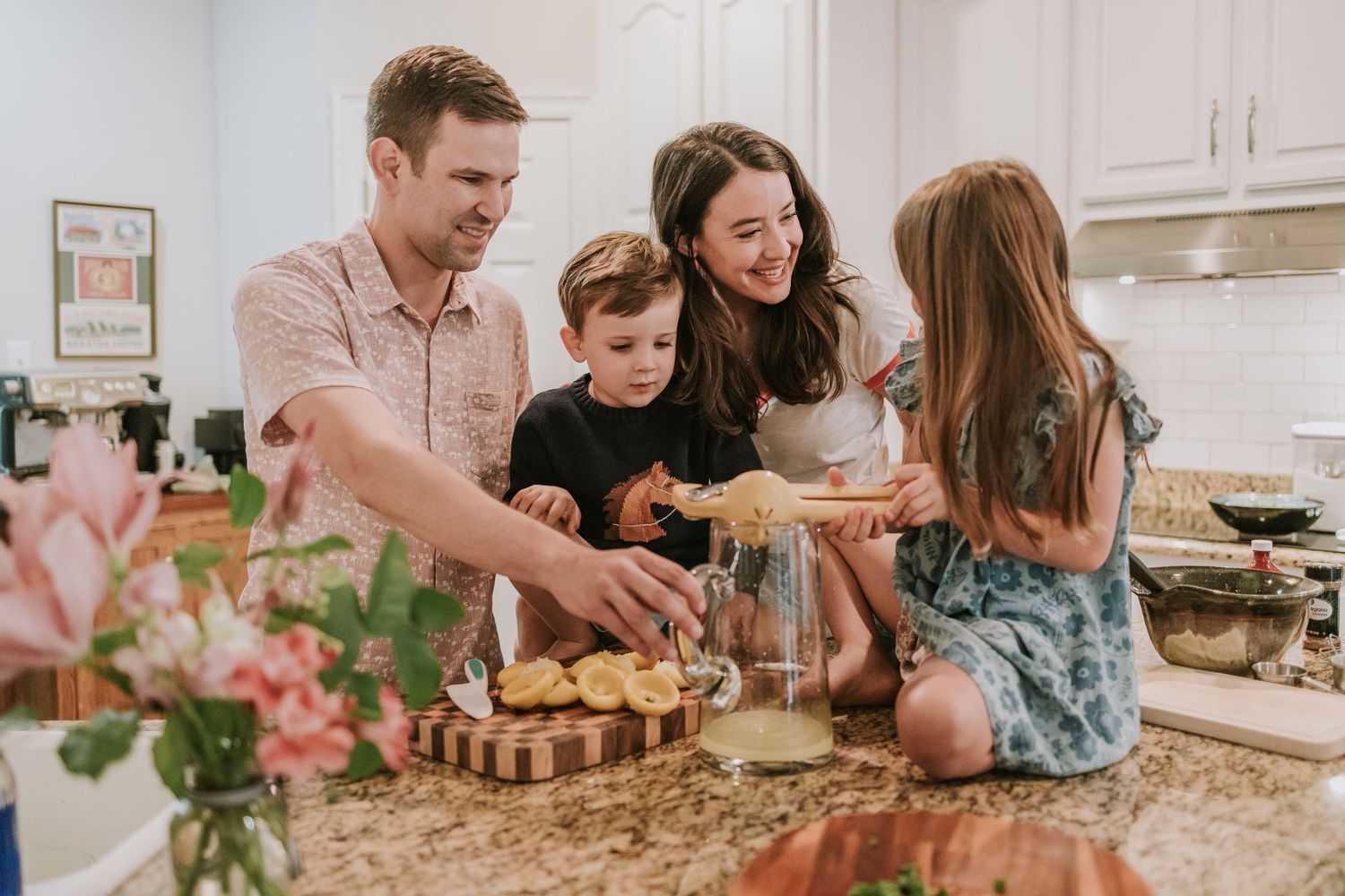 Family gathered around kitchen counter preparing food together with fresh flowers on display in foreground.