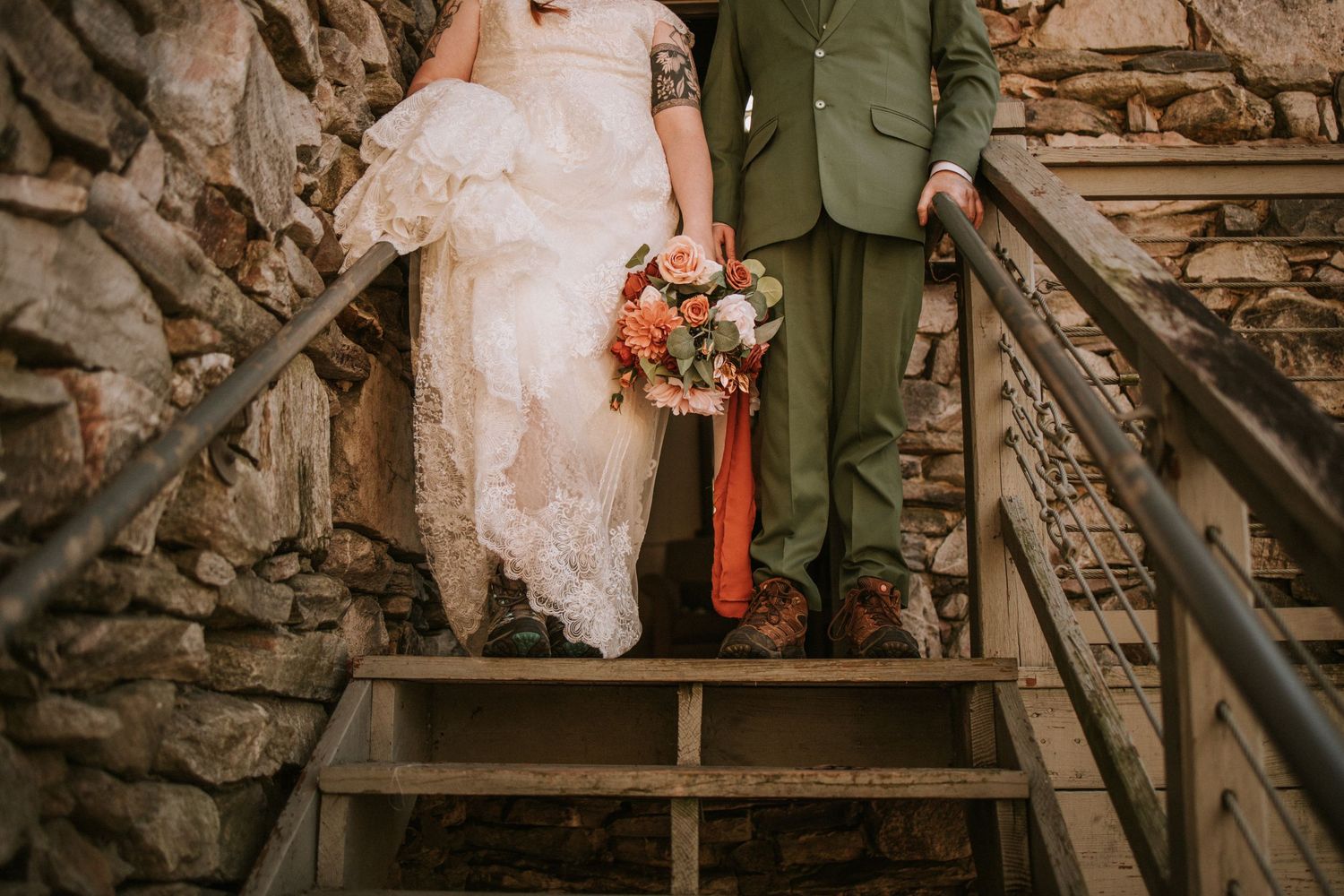 Close-up view of bride in white dress and groom in green suit descending rustic stone staircase at wedding.