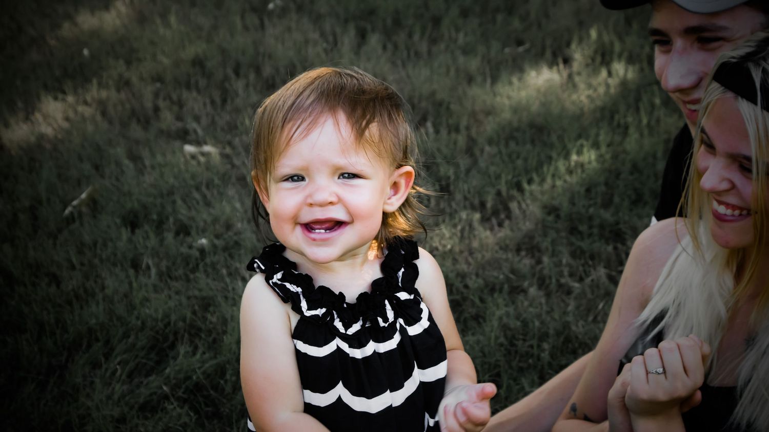 Baby girl smiling with parents beside her during outdoor family photo session