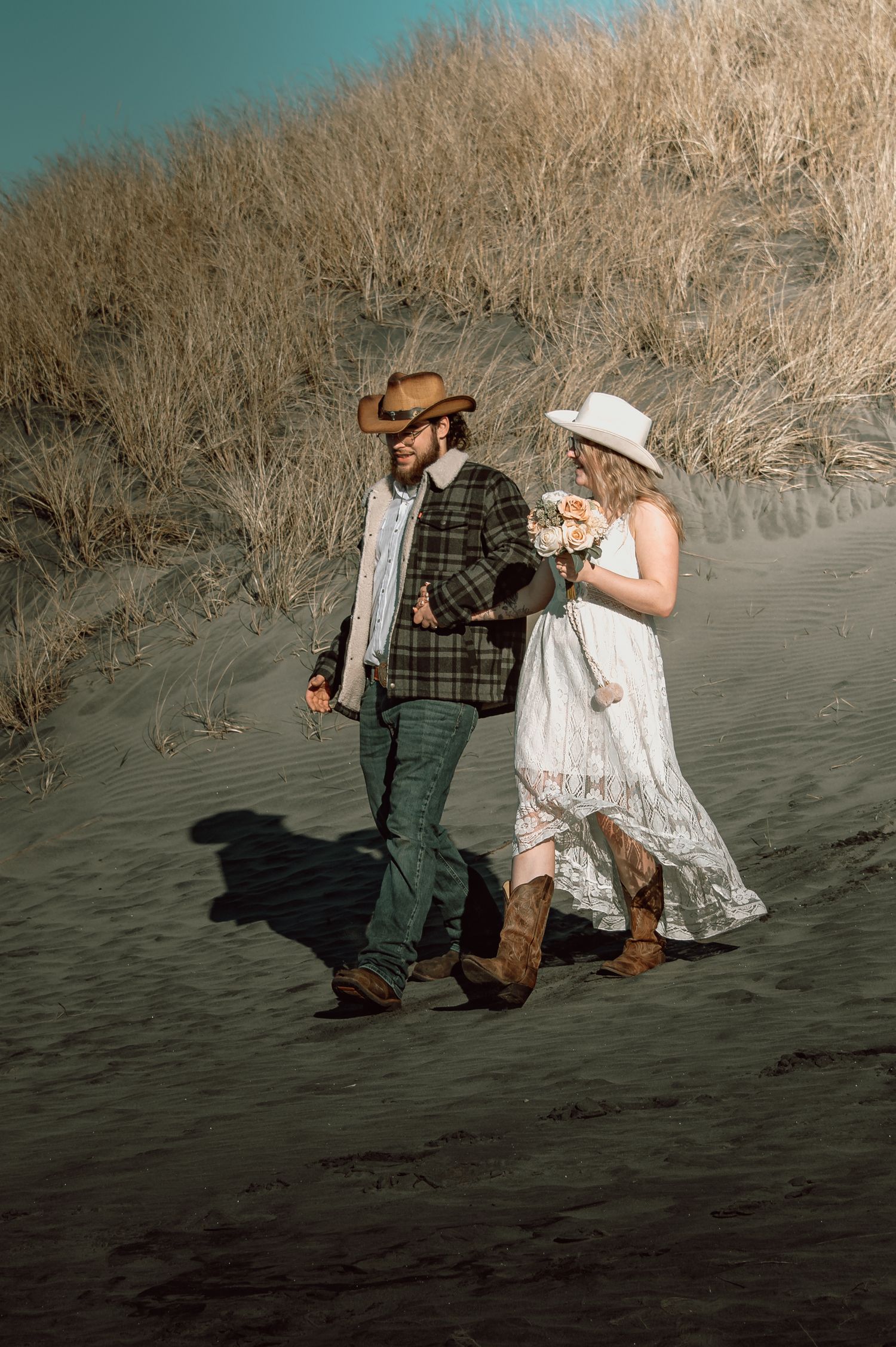 Couple in western attire walking on sandy beach dunes with dry coastal grasses in the background.