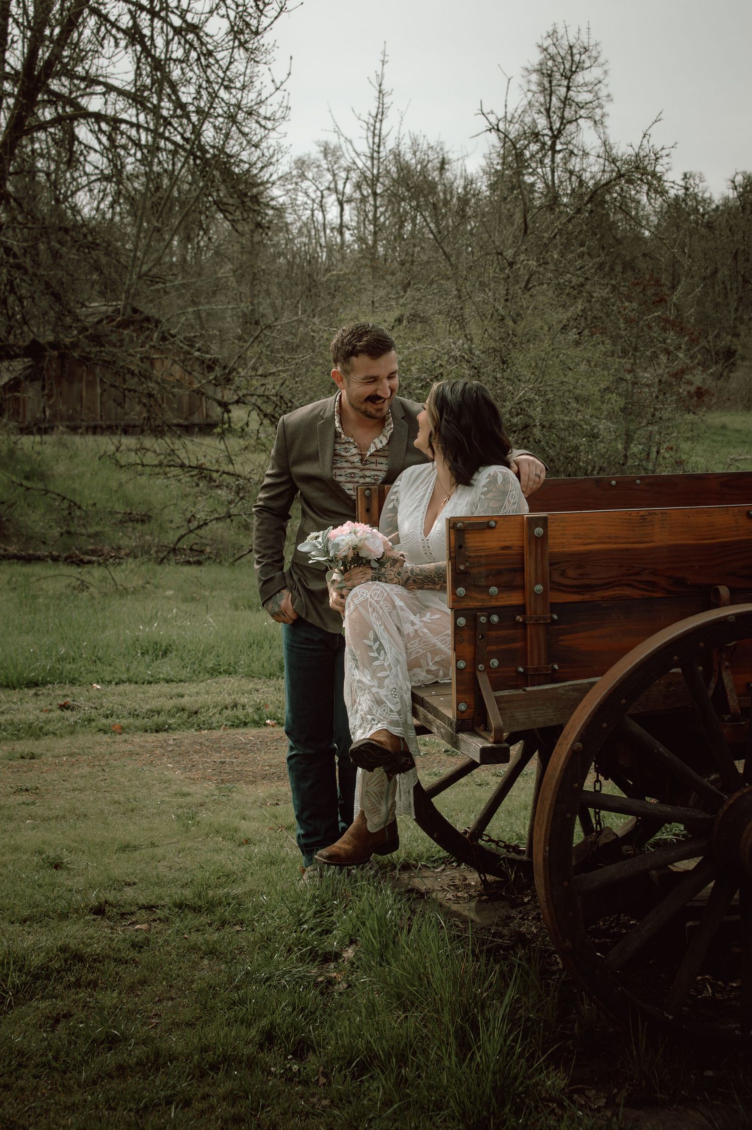 Groom in olive suit gazes at bride seated in vintage wooden wagon holding pink bouquet in outdoor setting.