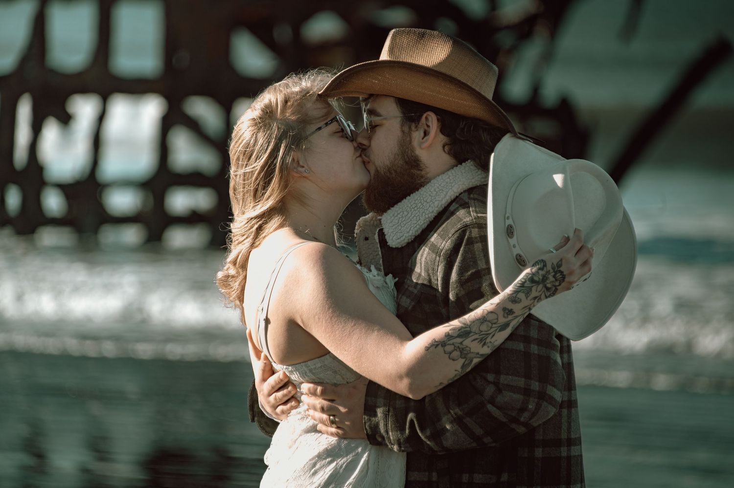 Couple sharing a romantic kiss on the beach, man wearing cowboy hat and woman in white dress.