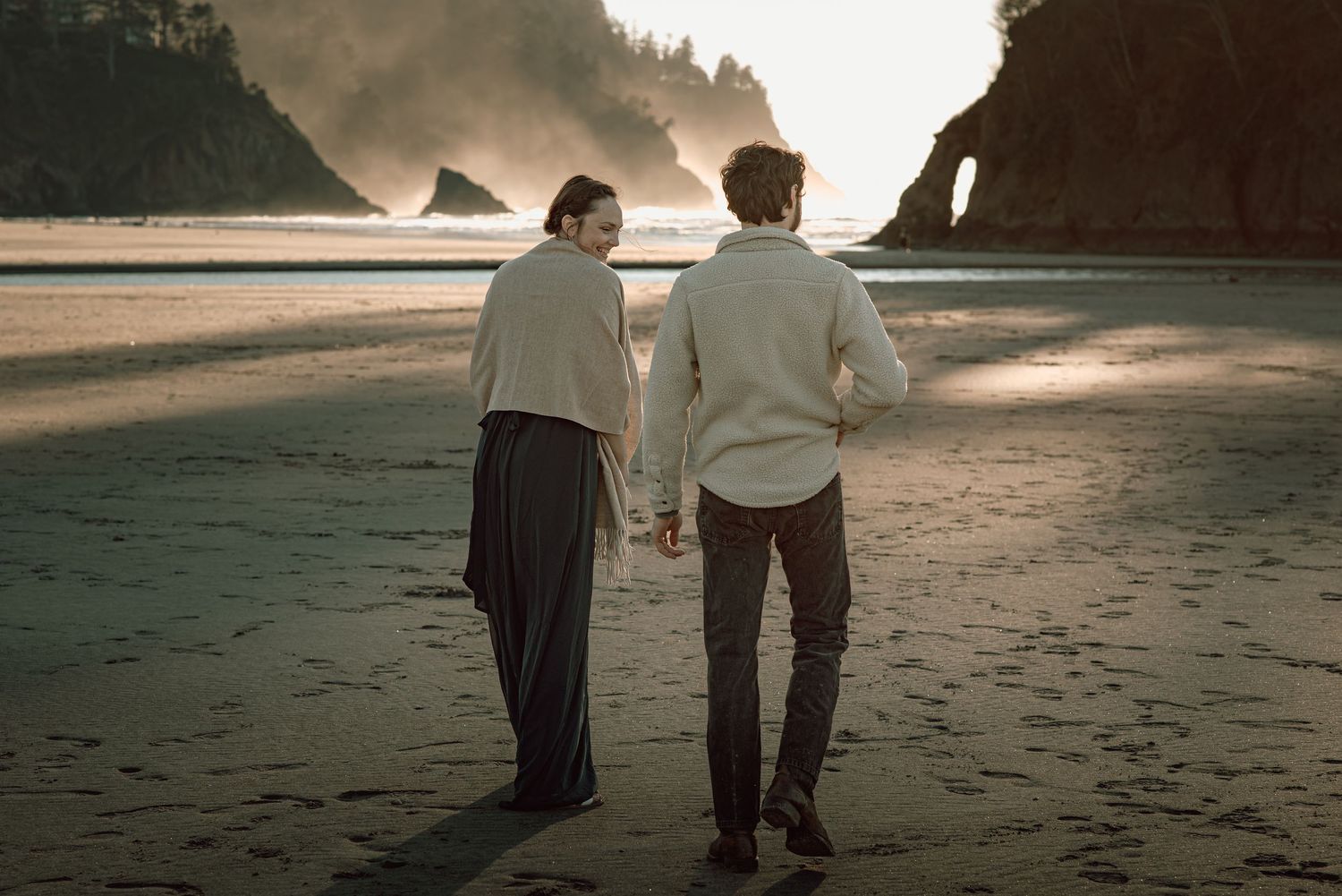 Couple walks hand in hand on a misty beach with dramatic sea stacks in the background.