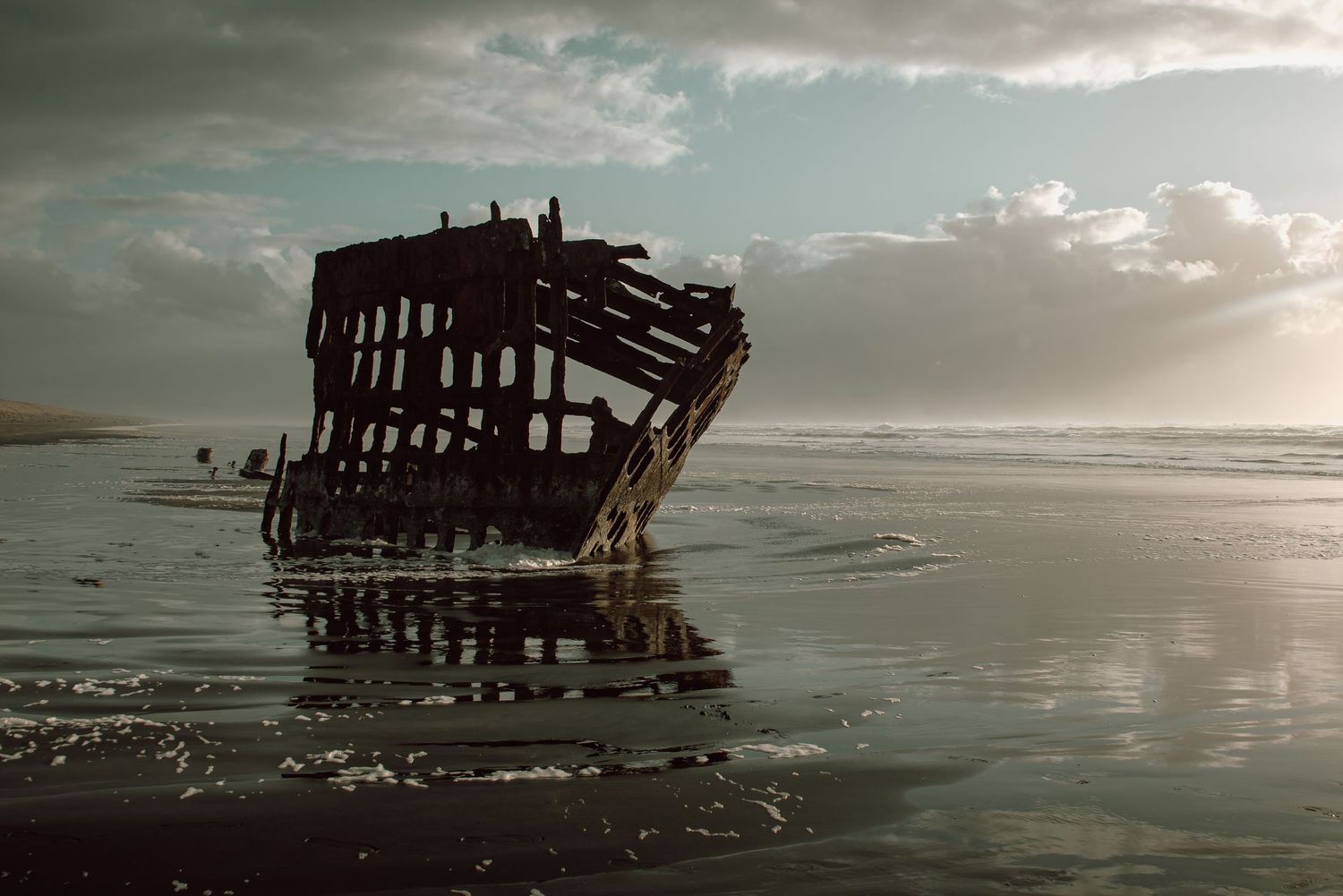 Weathered shipwreck ruins reflected in wet sand at low tide under a dramatic cloudy sky.