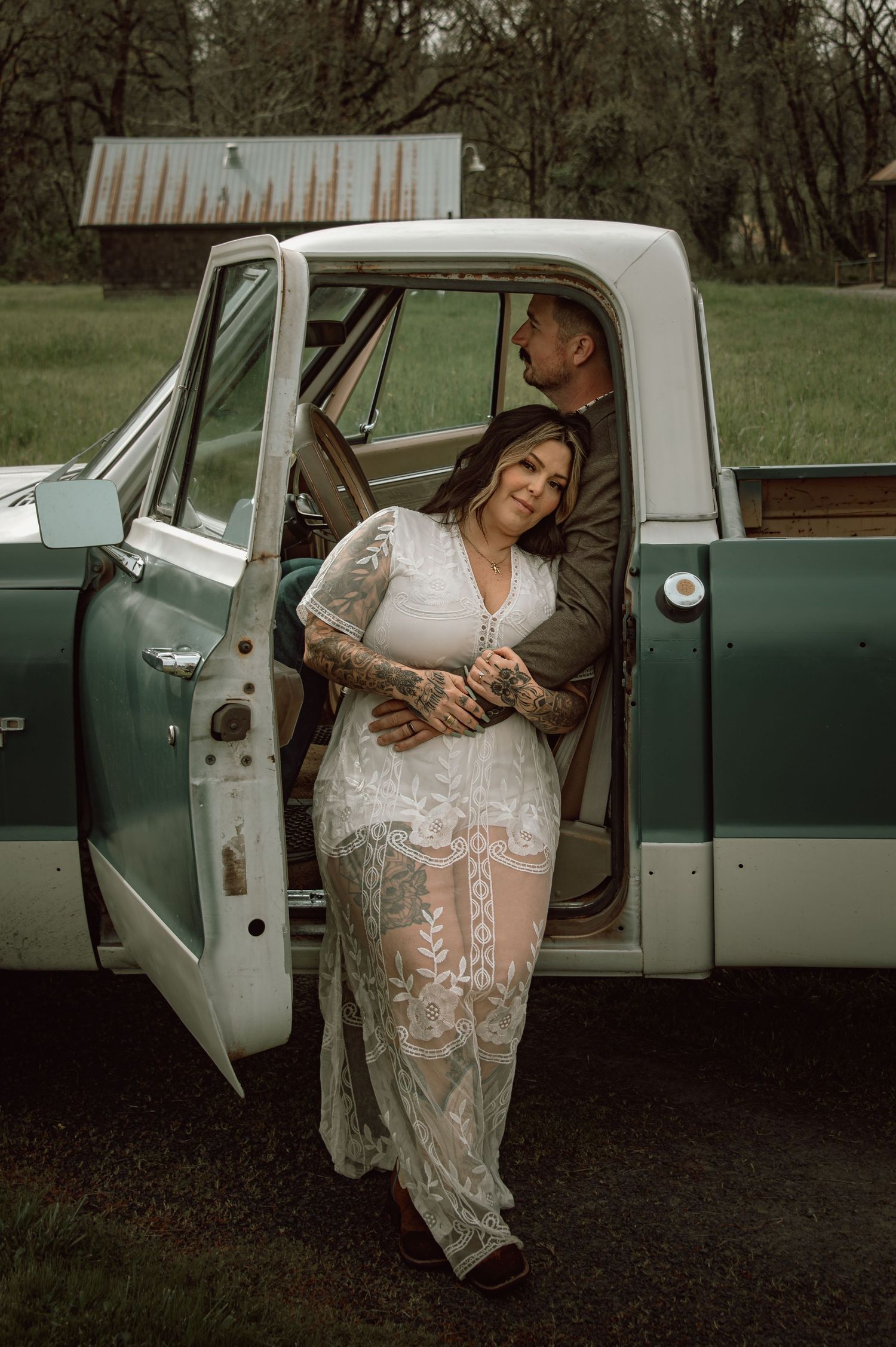 Bride in sheer lace dress leans against vintage teal pickup truck with groom in olive blazer.
