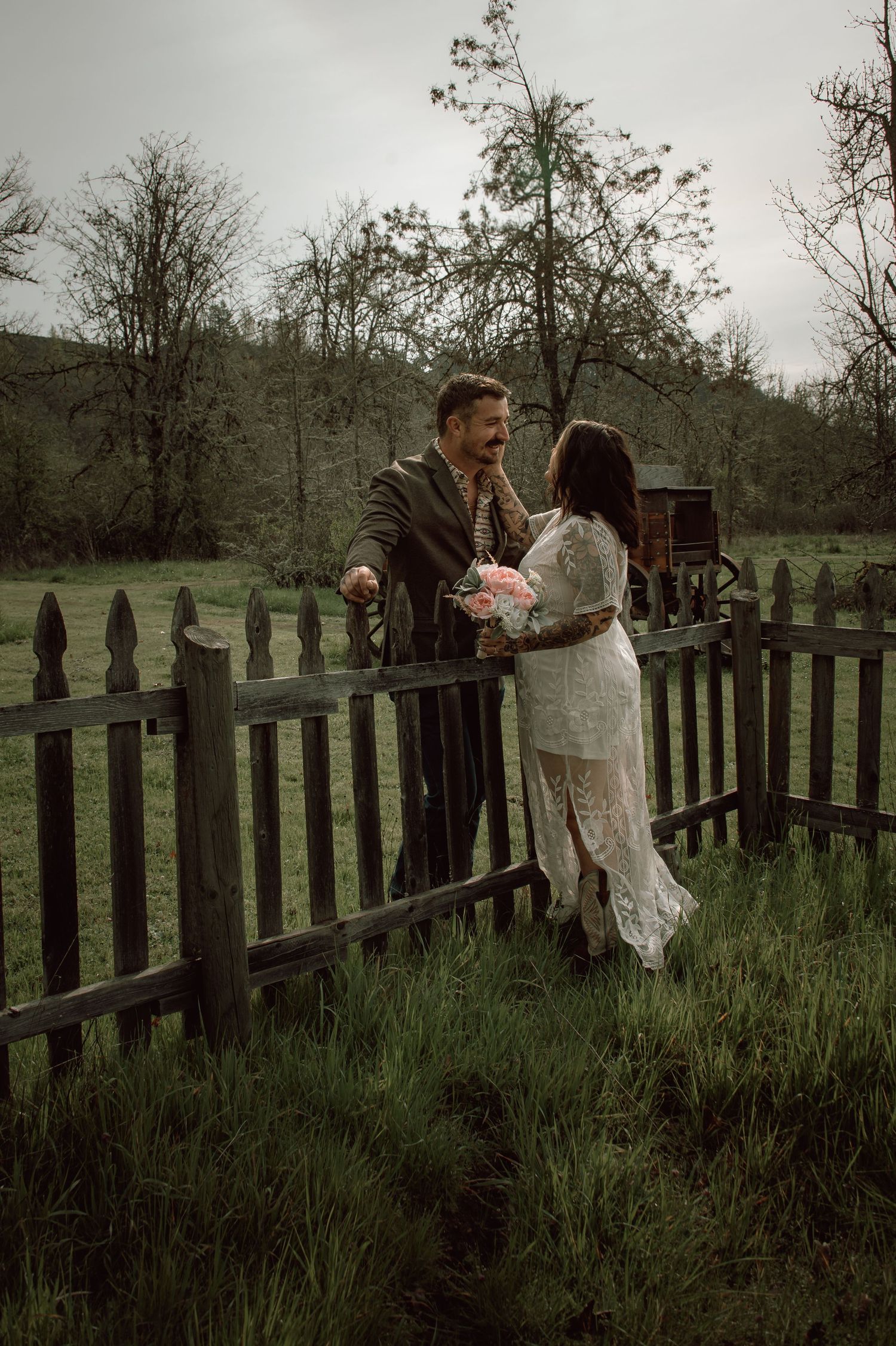 Couple sharing a joyful moment by a weathered wooden fence in a lush green rural farm setting.