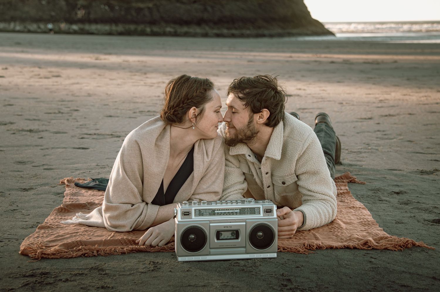 Couple sharing a kiss lying on a blanket on the beach with a vintage boombox nearby.