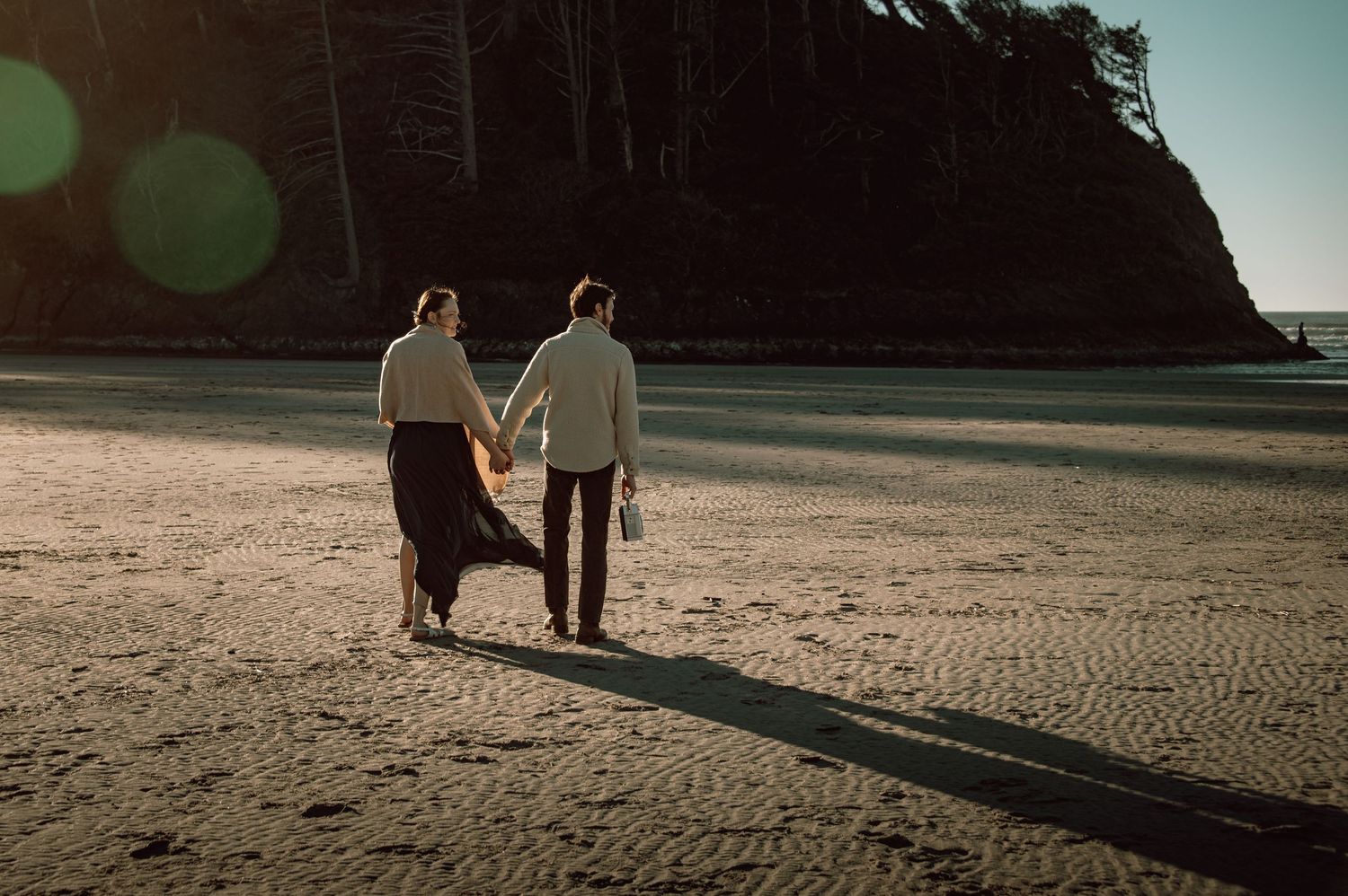 Couple holding hands walking on a sandy beach at sunset with dramatic coastal cliffs in the background.