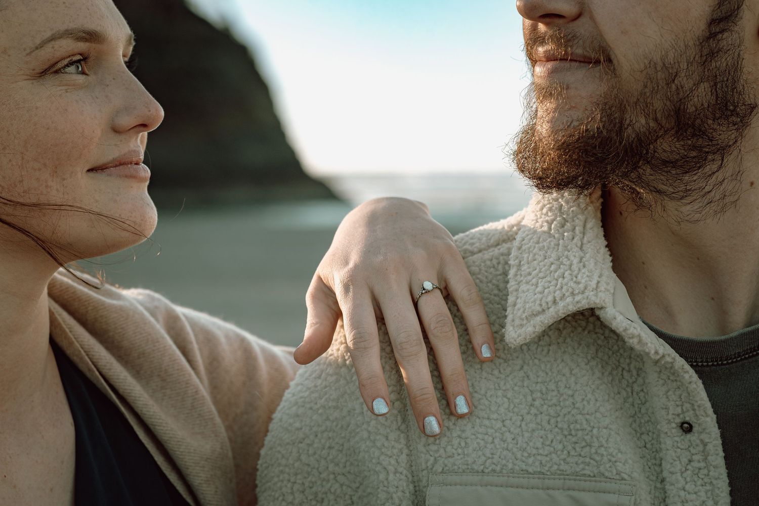 Close-up of woman showing diamond engagement ring on hand resting on bearded mans shoulder.