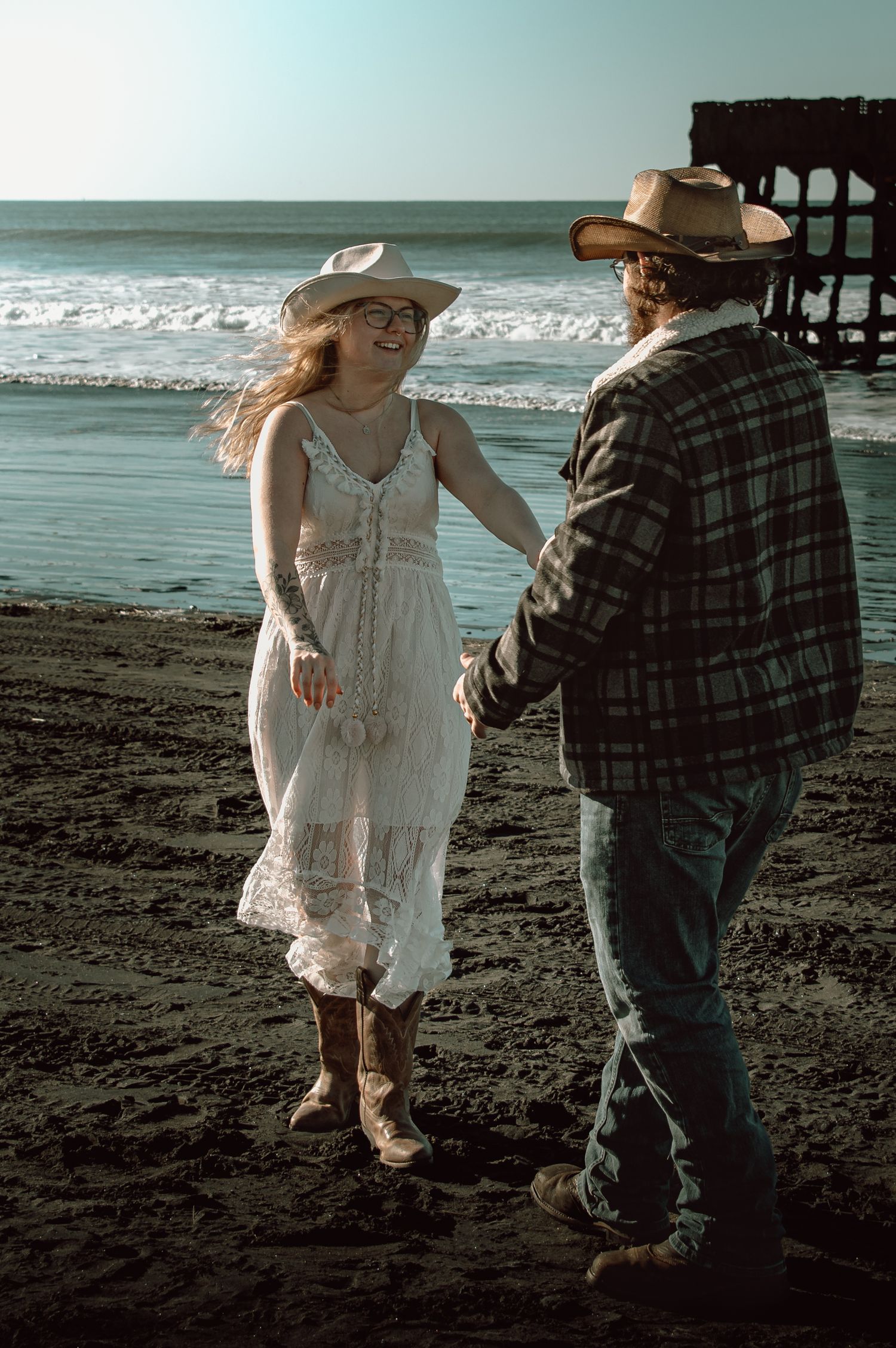Couple in cowboy hats hold hands and laugh together on a scenic ocean beach.