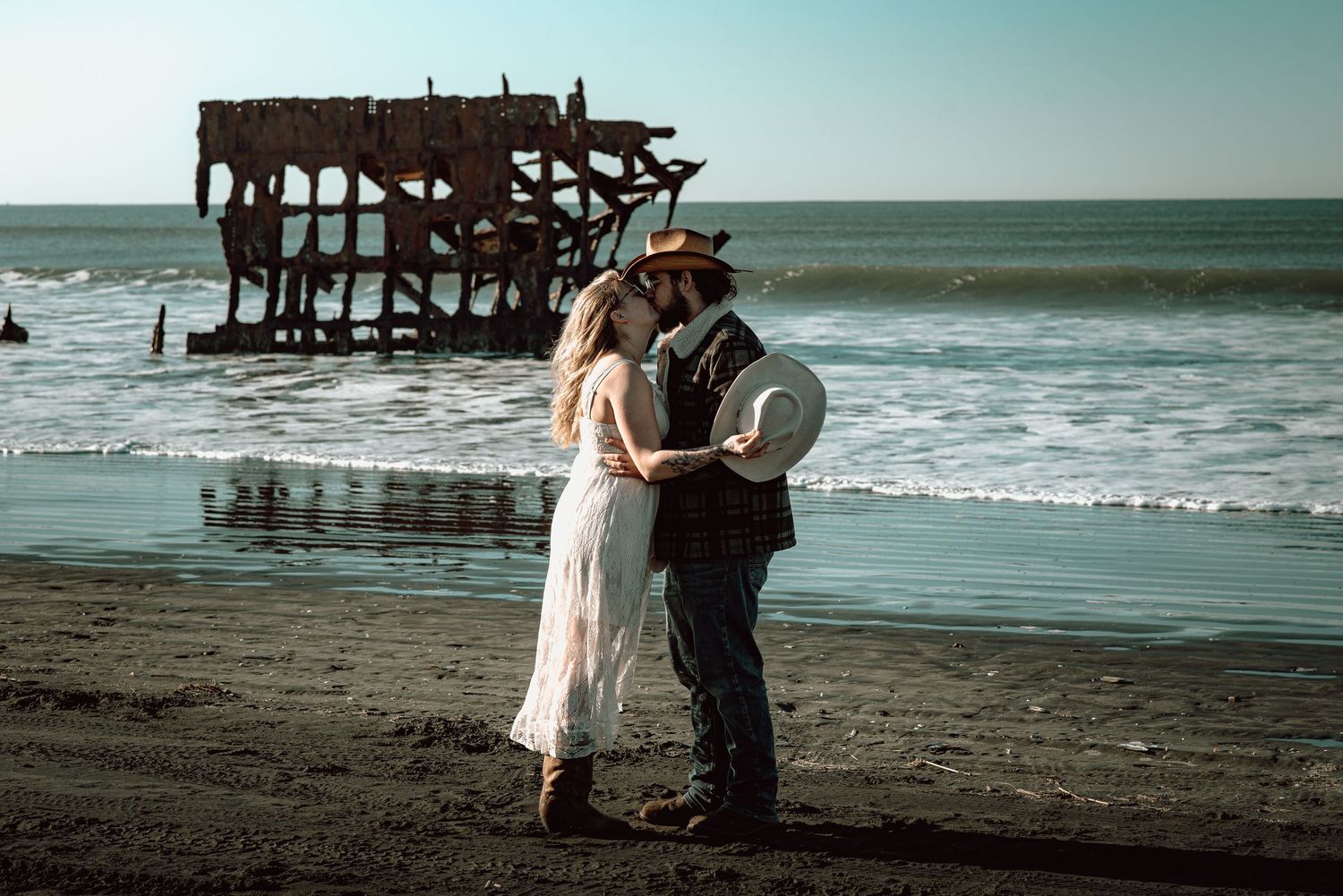 Couple sharing a romantic moment on a dark sand beach with a historic shipwreck in the ocean background.