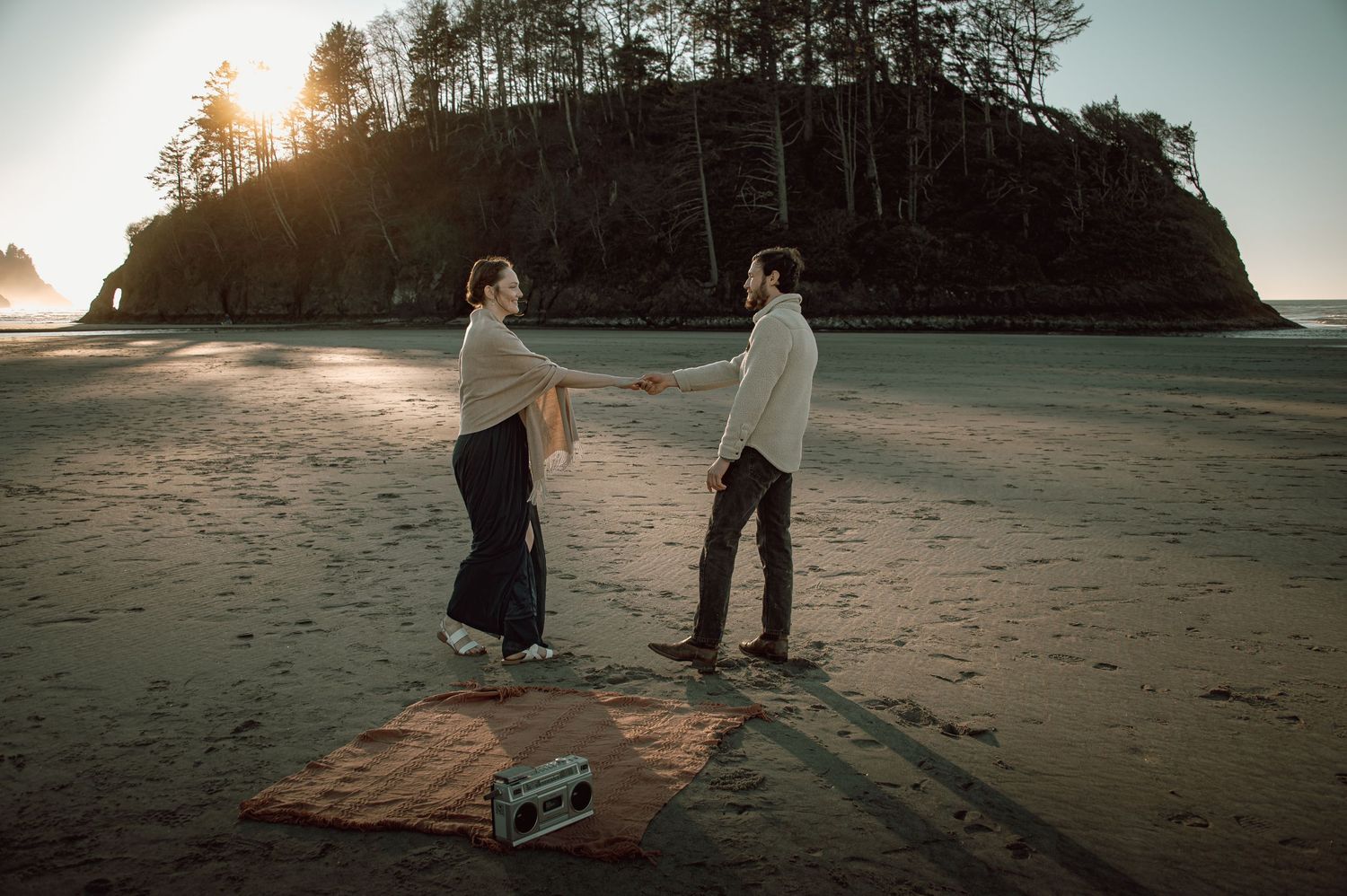 Couple holding hands facing each other on beach with vintage boombox and sea stack backdrop.