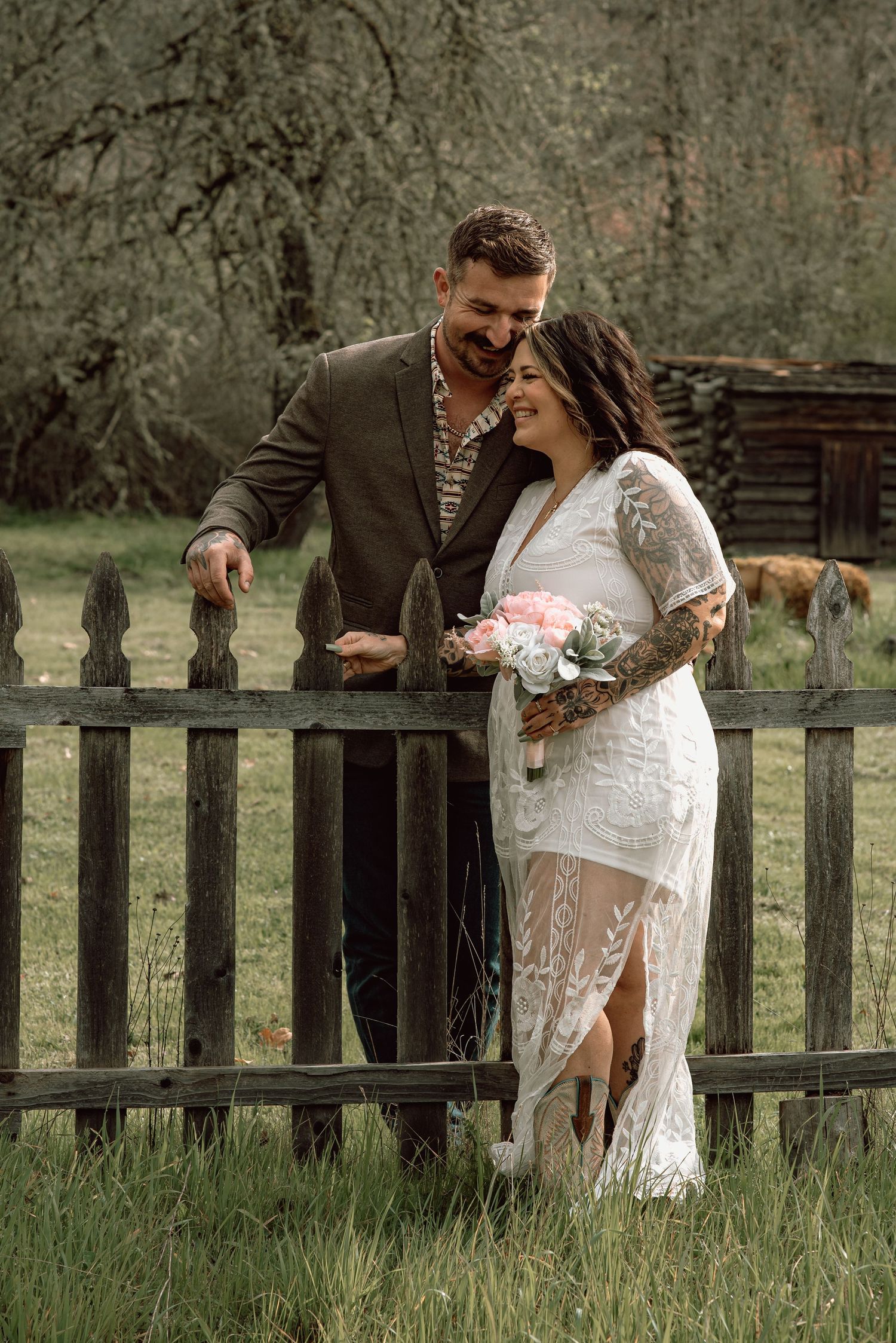 Smiling tattooed couple walks along rustic wooden fence, bride holds pink bouquet in lace dress.
