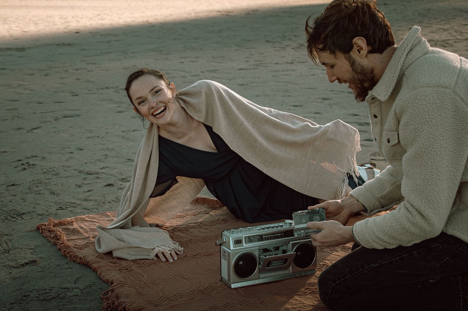 Woman laughing on beach blanket while man sets up a vintage boombox during golden hour.