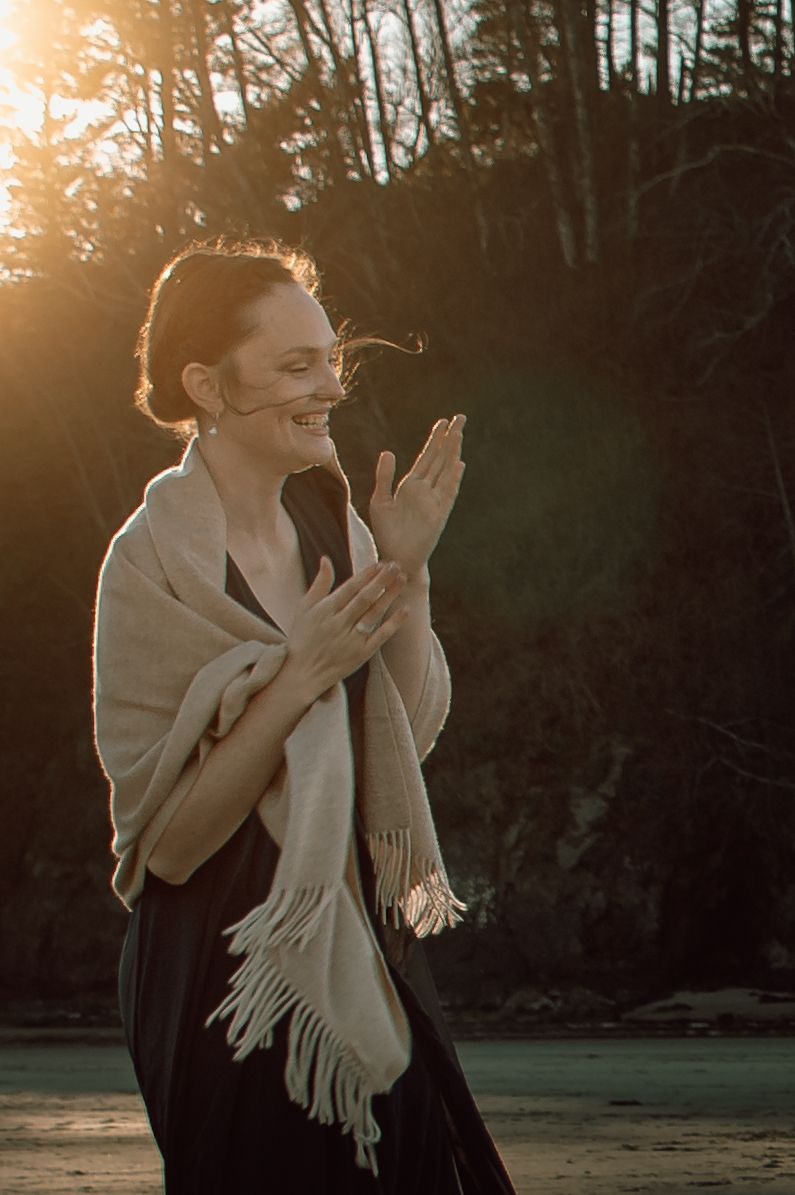 Happy woman wrapped in fringe shawl clapping at beach sunset.