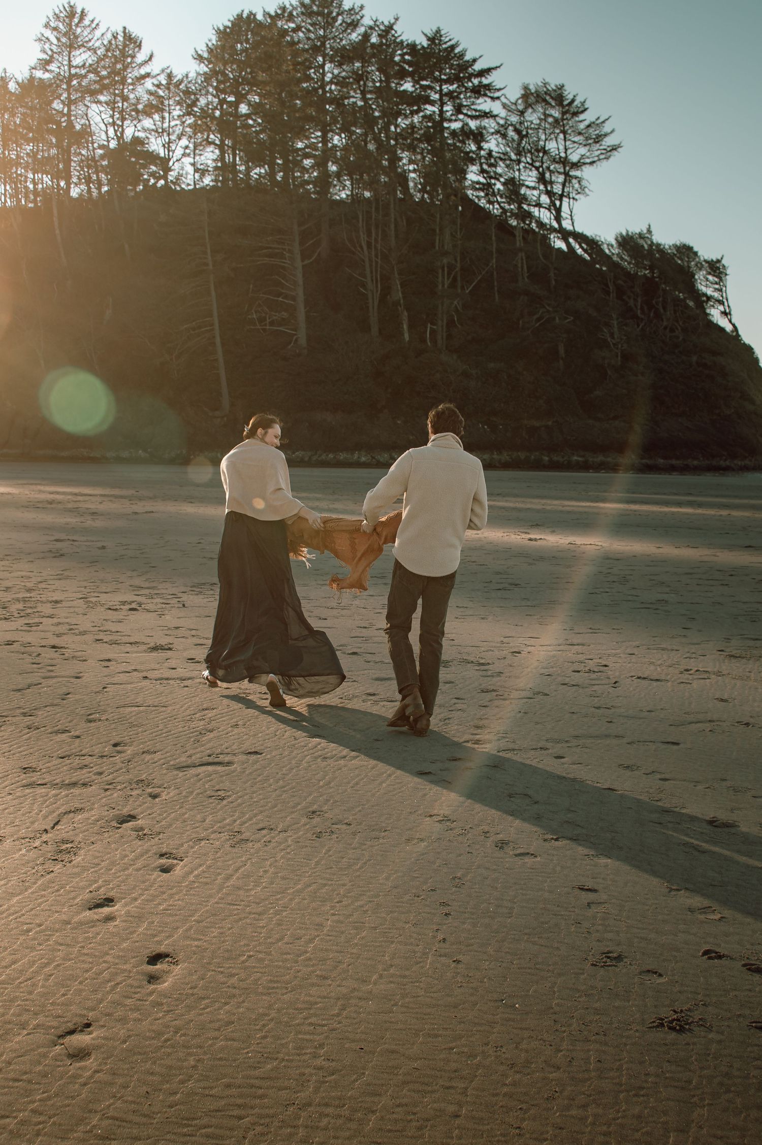 Couple walking hand in hand on a sunlit beach carrying a blanket with trees in background.