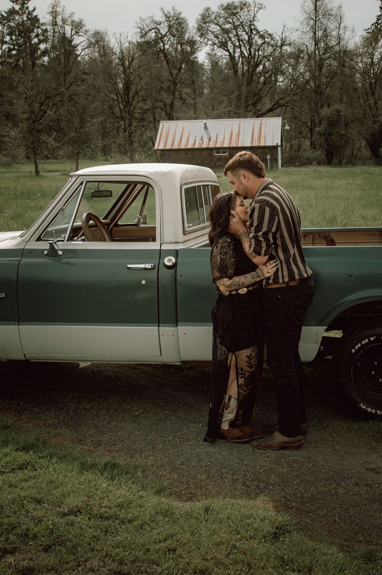 Couple sharing a romantic kiss leaning against a classic teal pickup truck in a green countryside field.
