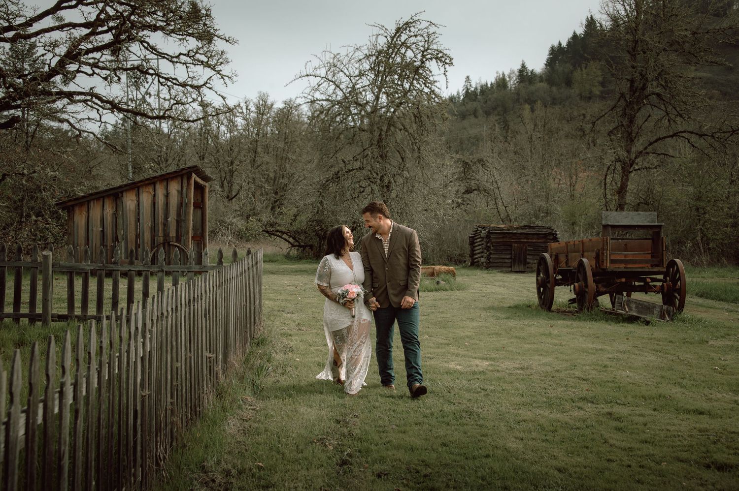 Bride and groom walk together along rustic wooden fence with vintage wagon and bare trees in background.