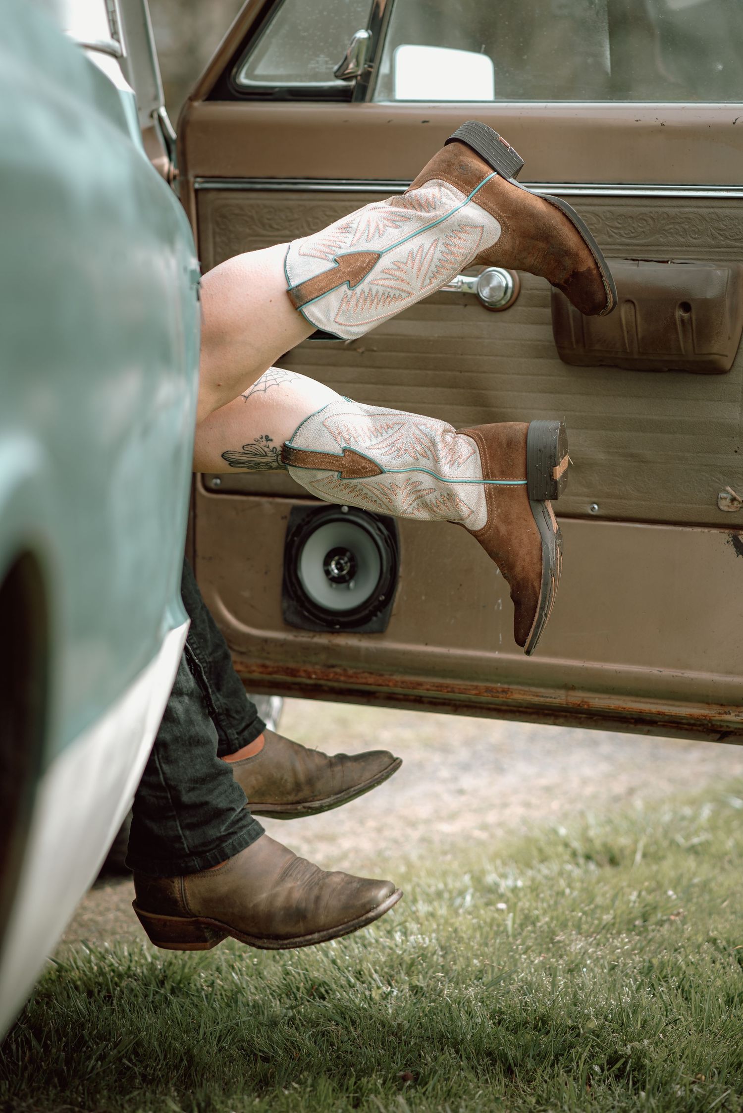 Two people sitting in a vintage car doorway wearing cowboy boots, legs dangling outside the door.