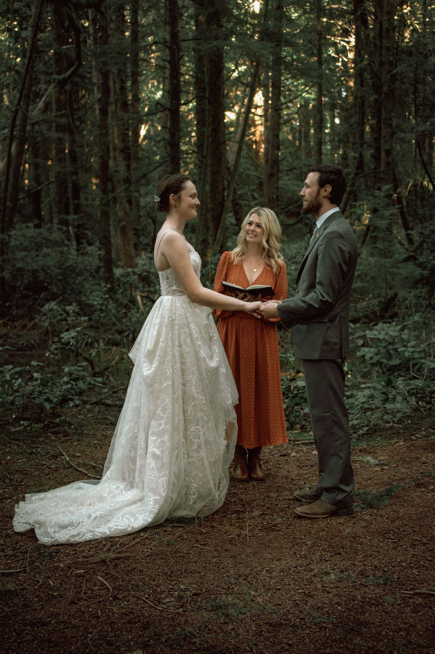 Bride and groom hold hands smiling during outdoor forest wedding ceremony with officiant in orange dress.
