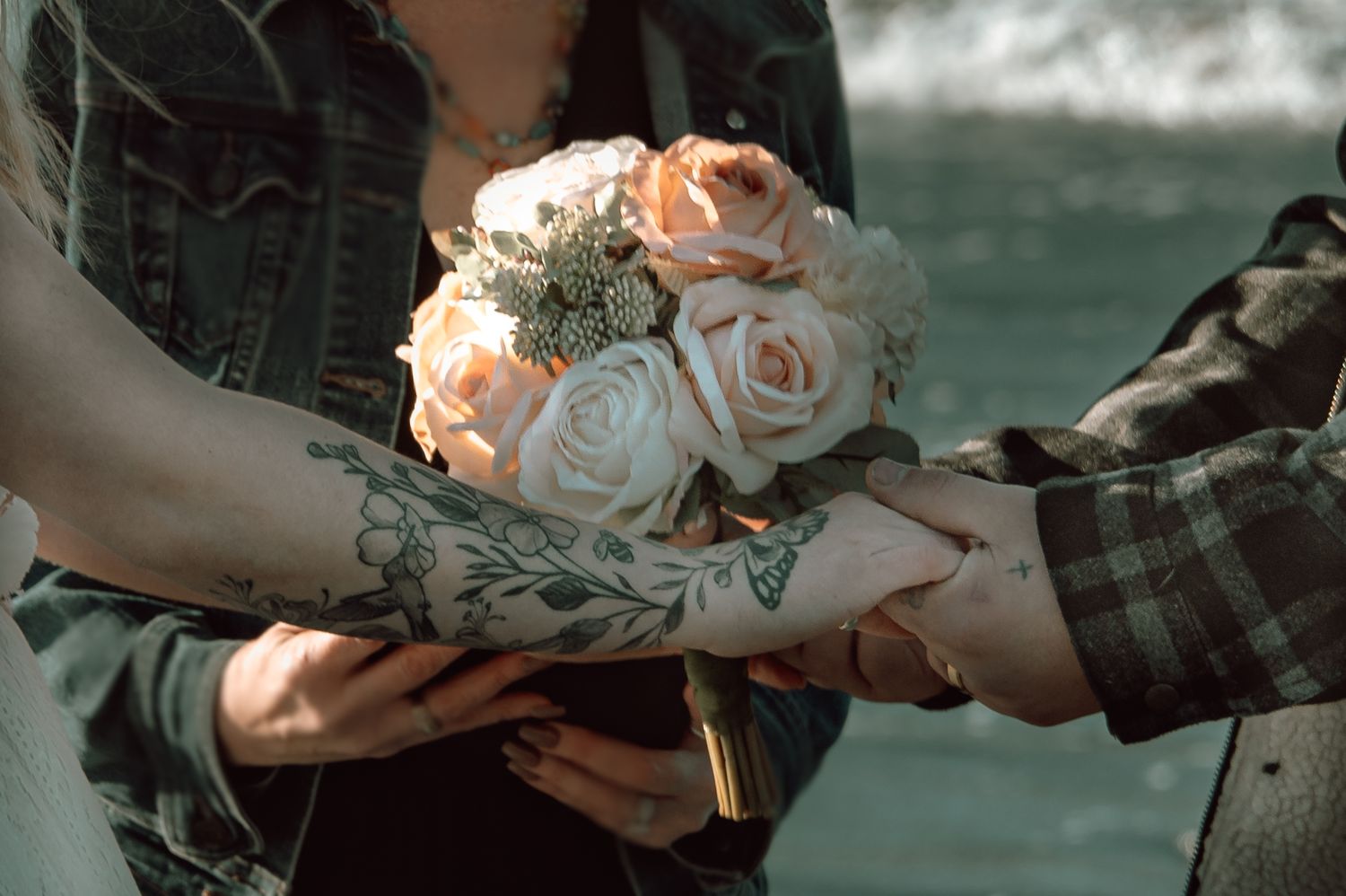 Close-up of tattooed hands holding delicate peach rose bouquet near ocean shore.