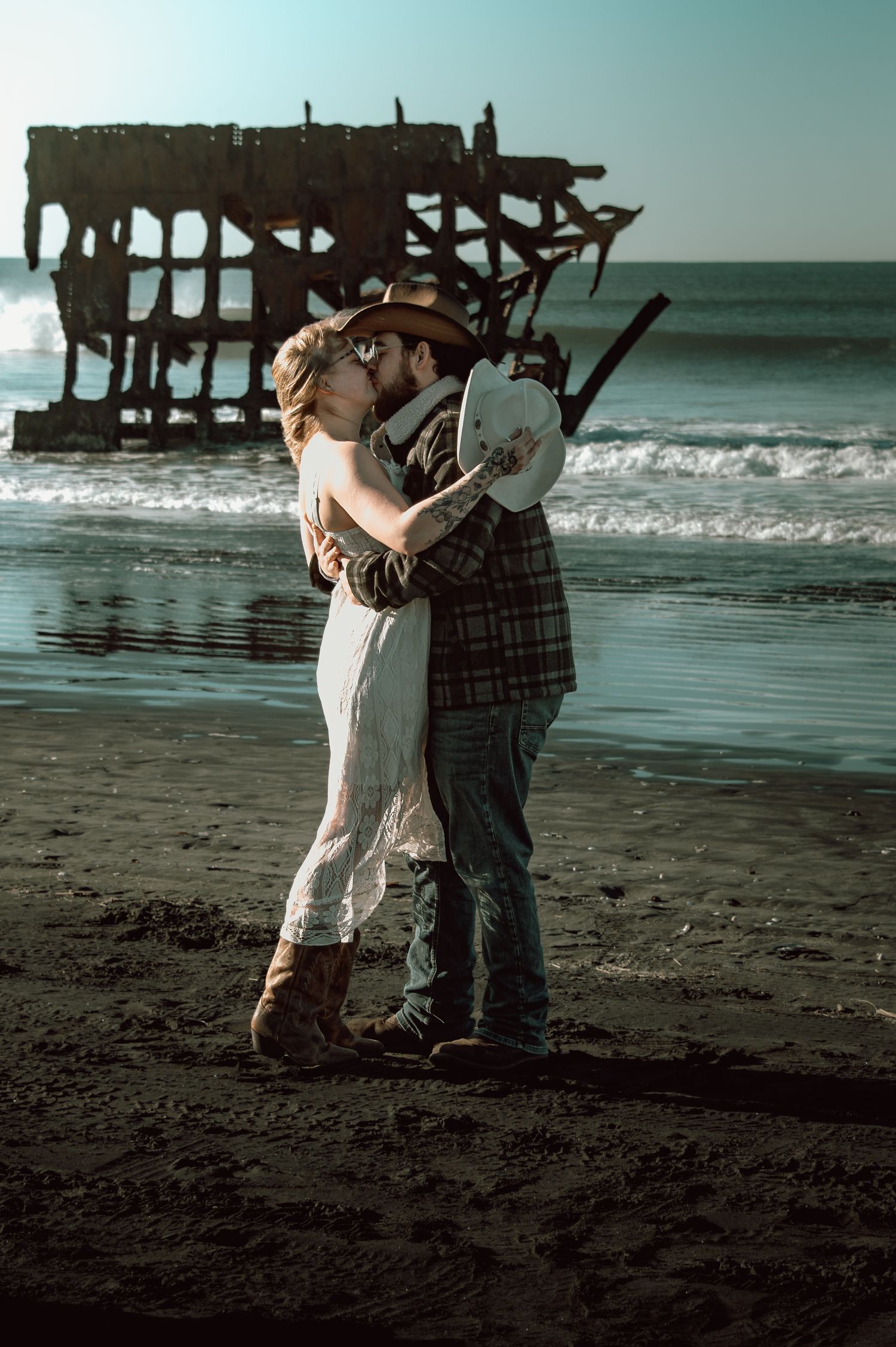 Couple embracing on a dark sand beach with a rustic shipwreck and ocean waves in the background.