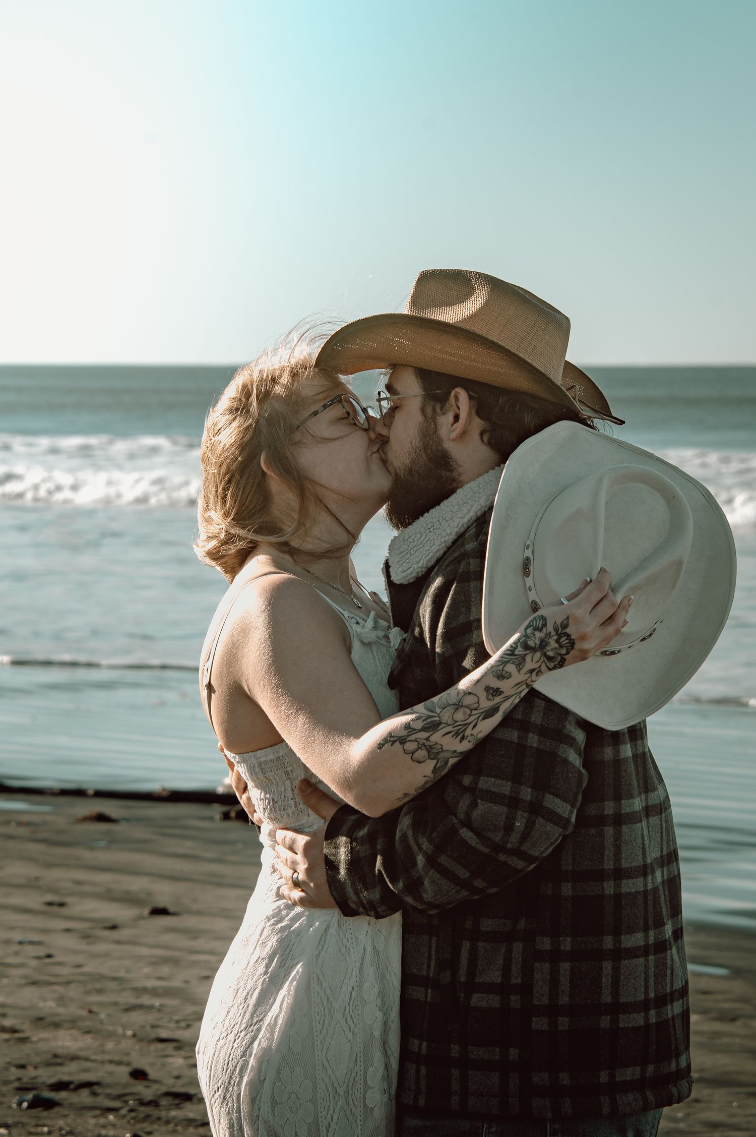 Couple sharing a passionate kiss on the beach, man wearing a cowboy hat and plaid jacket.
