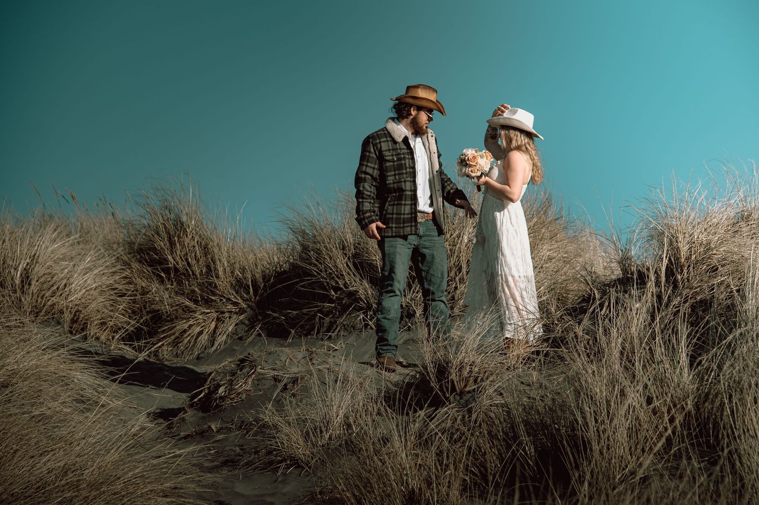 Couple in western attire standing among coastal sand dunes with dry grass under a teal sky.