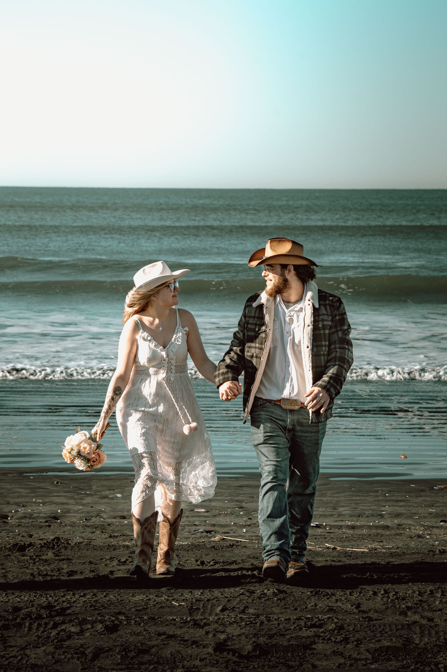 Bride and groom in cowboy hats walking hand in hand on a dark sandy beach with ocean waves behind them.