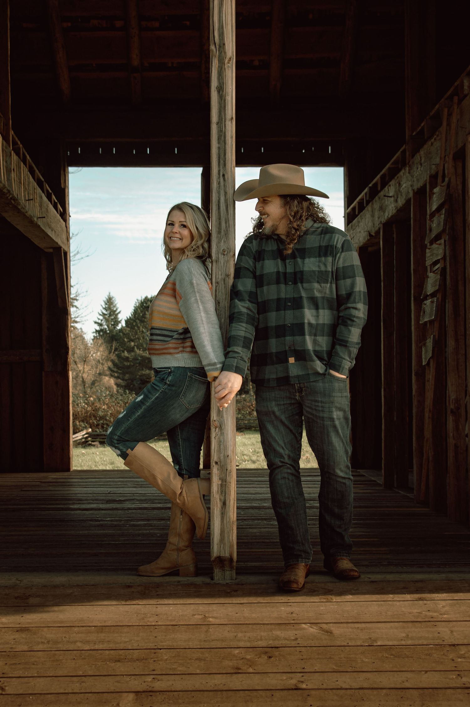Couple posing in rustic barn doorway wearing western flannel and cowboy hat with country backdrop.