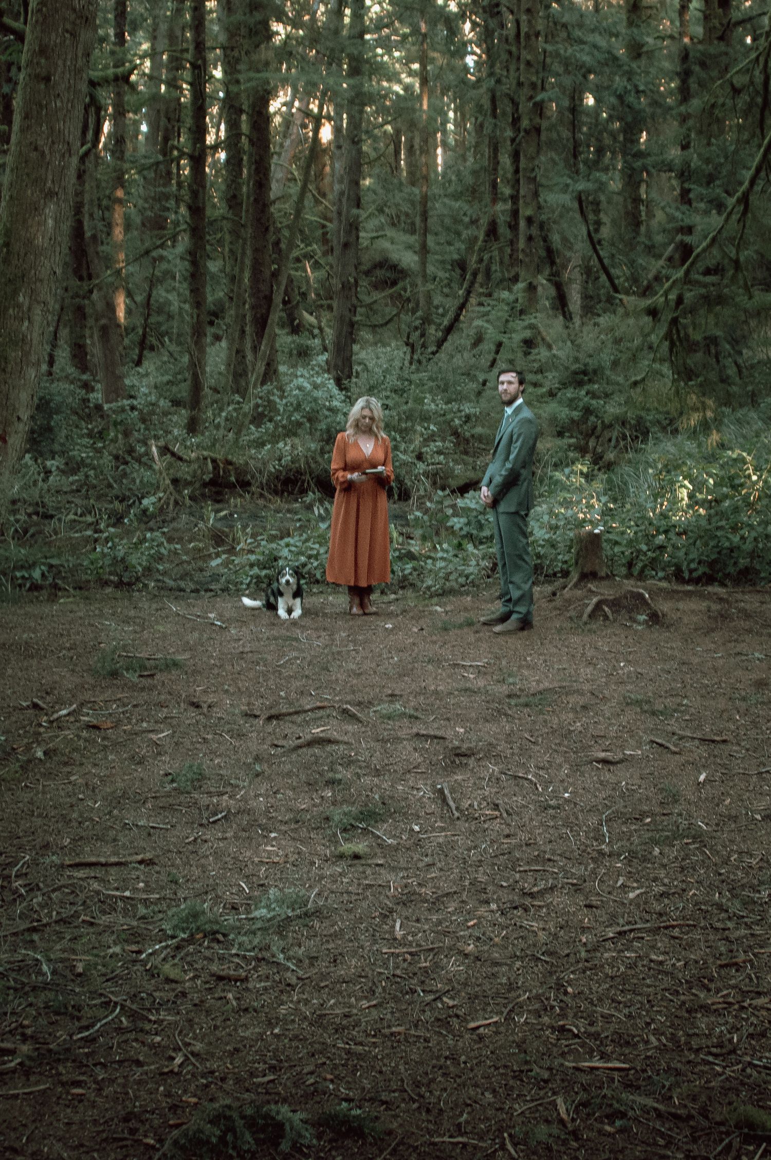 Couple stands with their dog in a serene forest clearing during outdoor wedding.