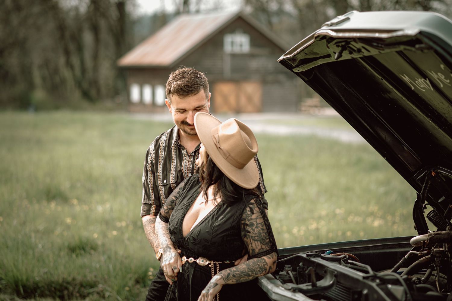 Tattooed couple looking under the hood of a vintage car on a rural farm property.