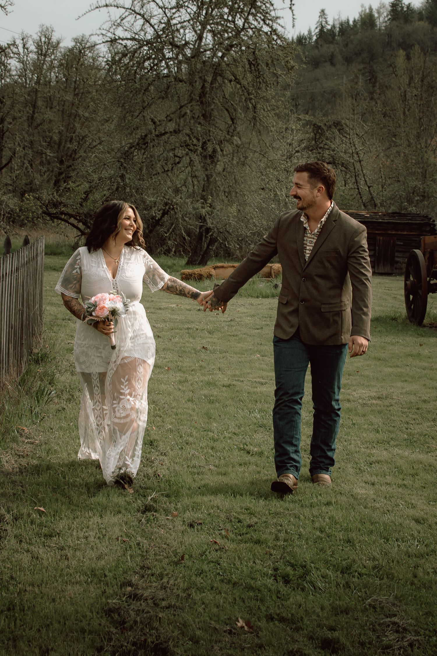 Laughing bride in lace gown holds pink bouquet while walking hand-in-hand with groom across green farm field.