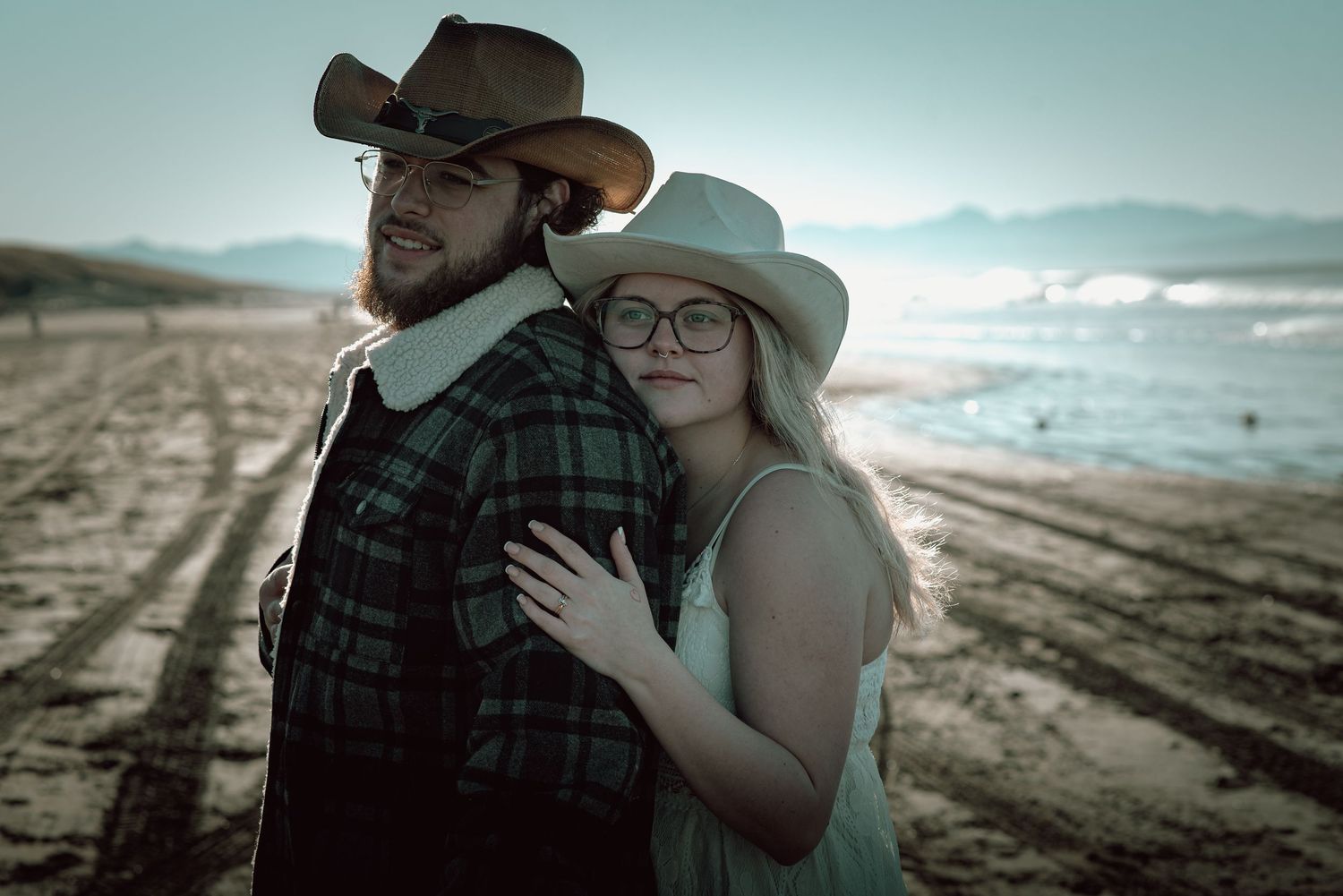 Western couple in cowboy hats sharing tender moment on sandy beach at golden hour.