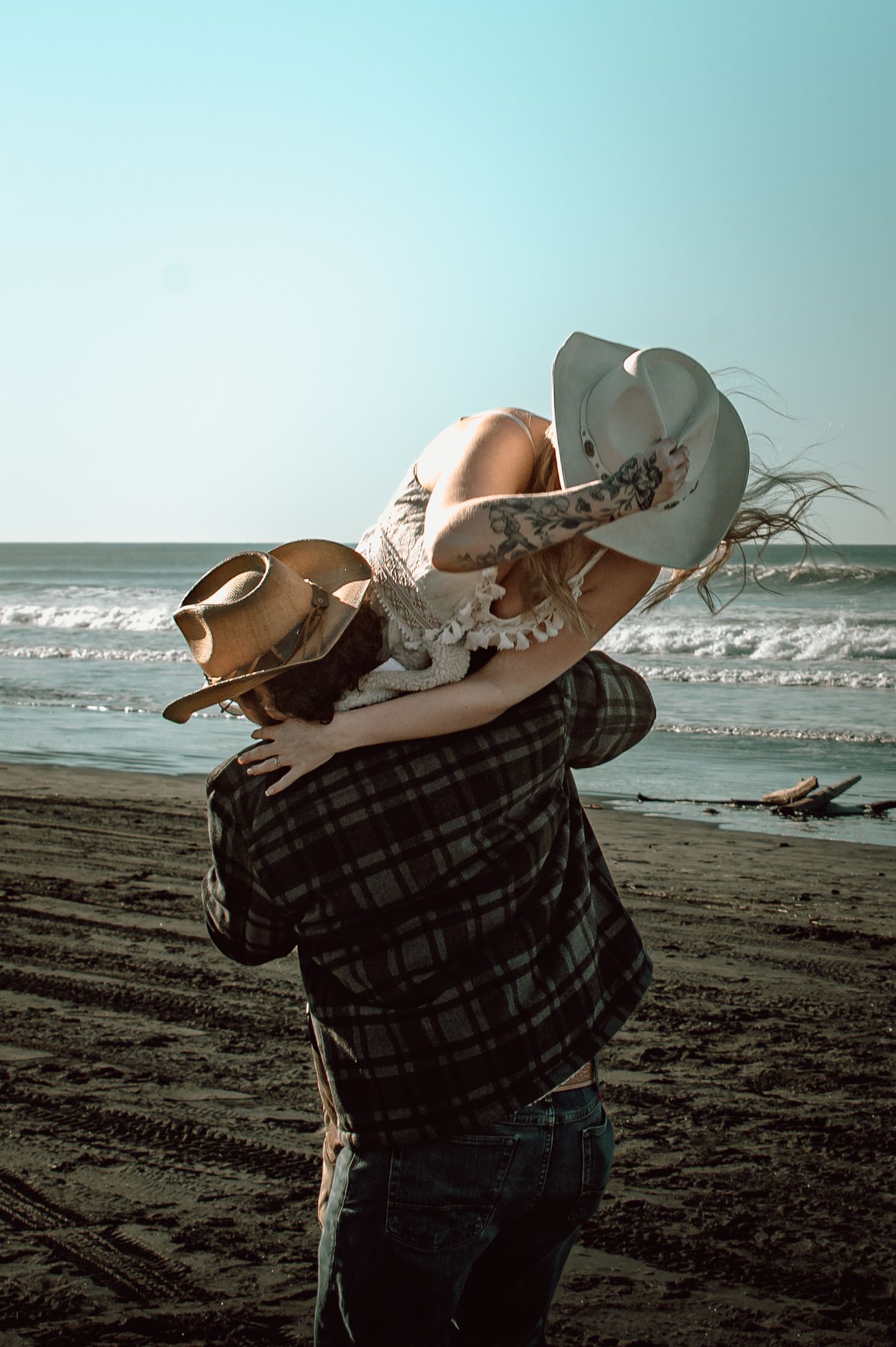 Couple in matching cowboy hats share a joyful embrace on a sunny beach by the ocean.