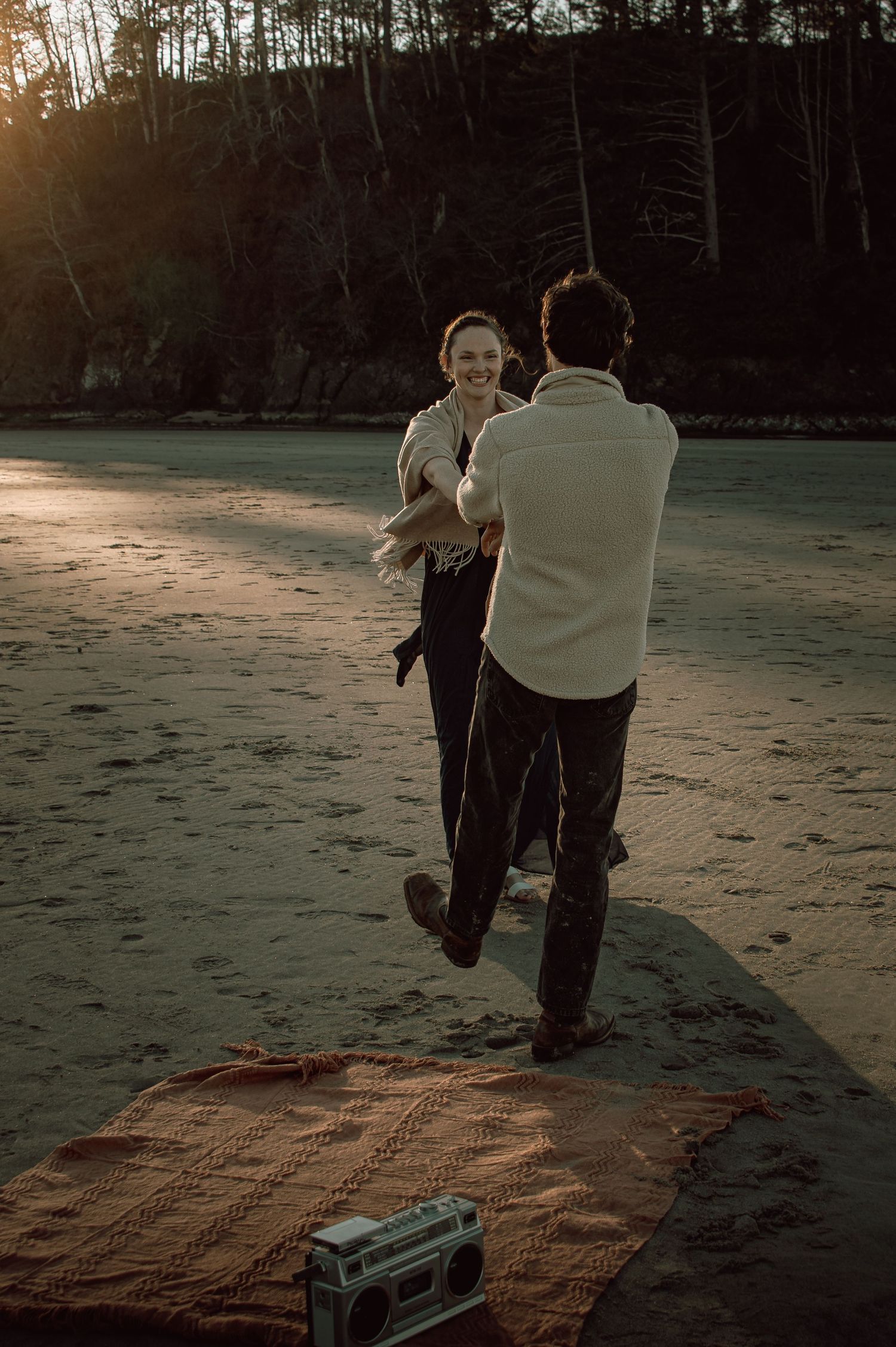 Smiling woman dancing with man on sandy beach at sunset with boombox on blanket nearby.