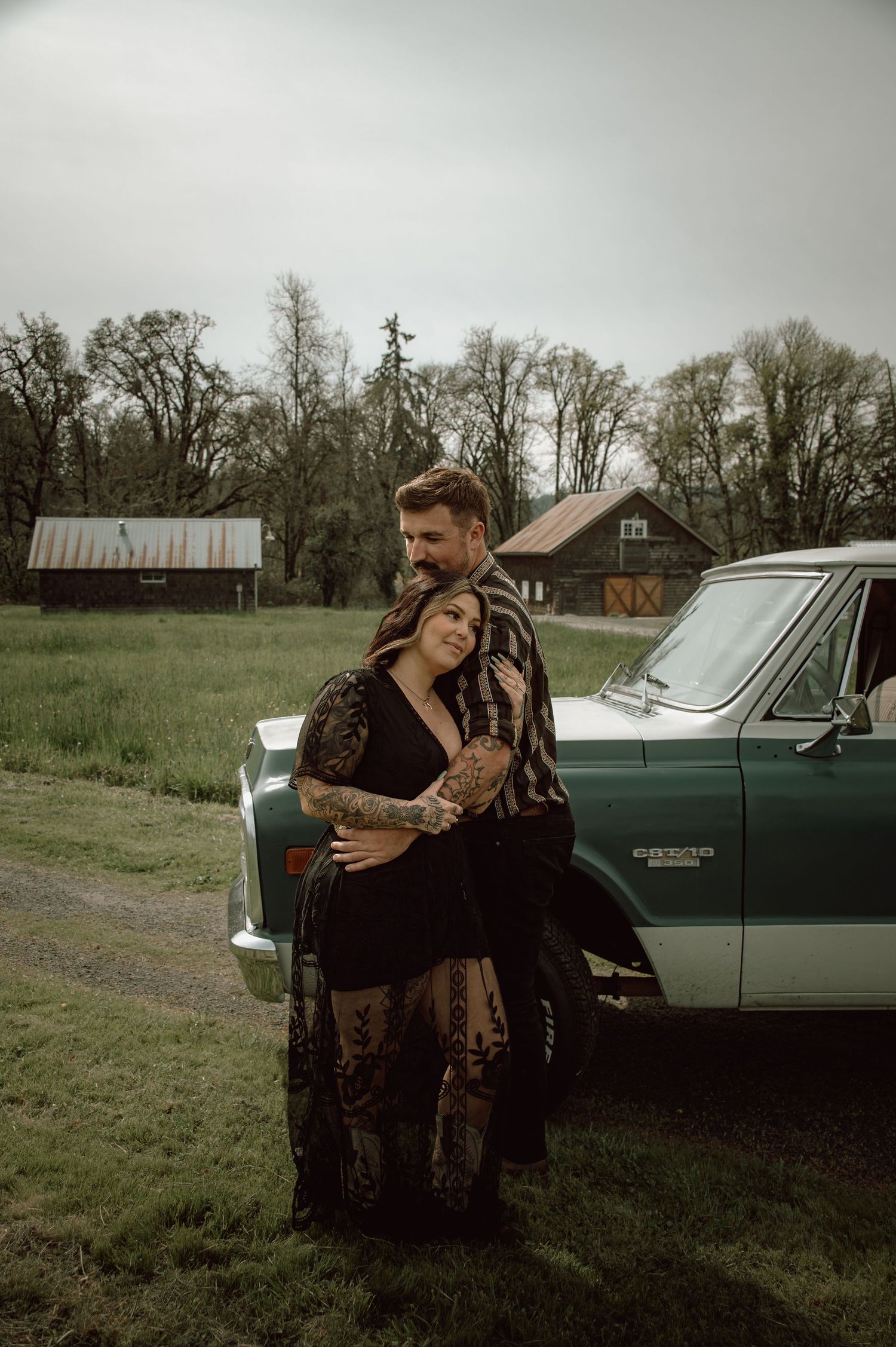 Tattooed couple posing together leaning against a classic teal truck in a green field.