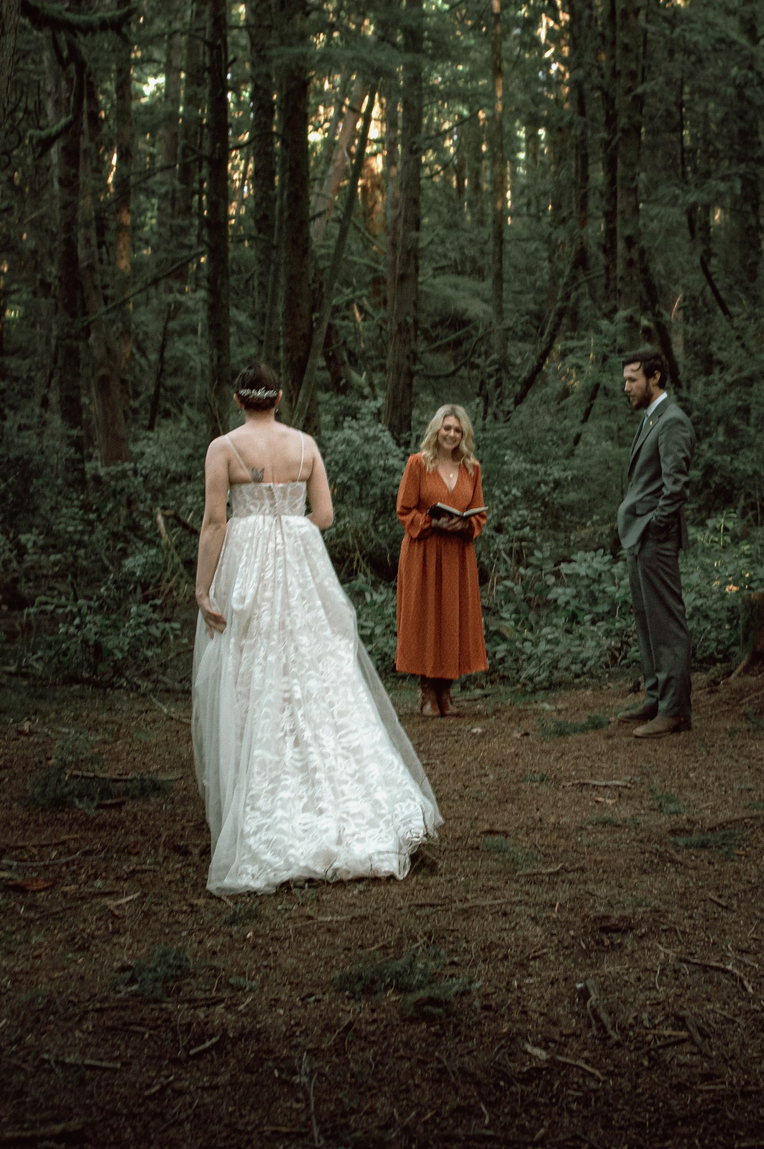 Bride in floral wedding gown faces officiant in rust dress during forest elopement ceremony.