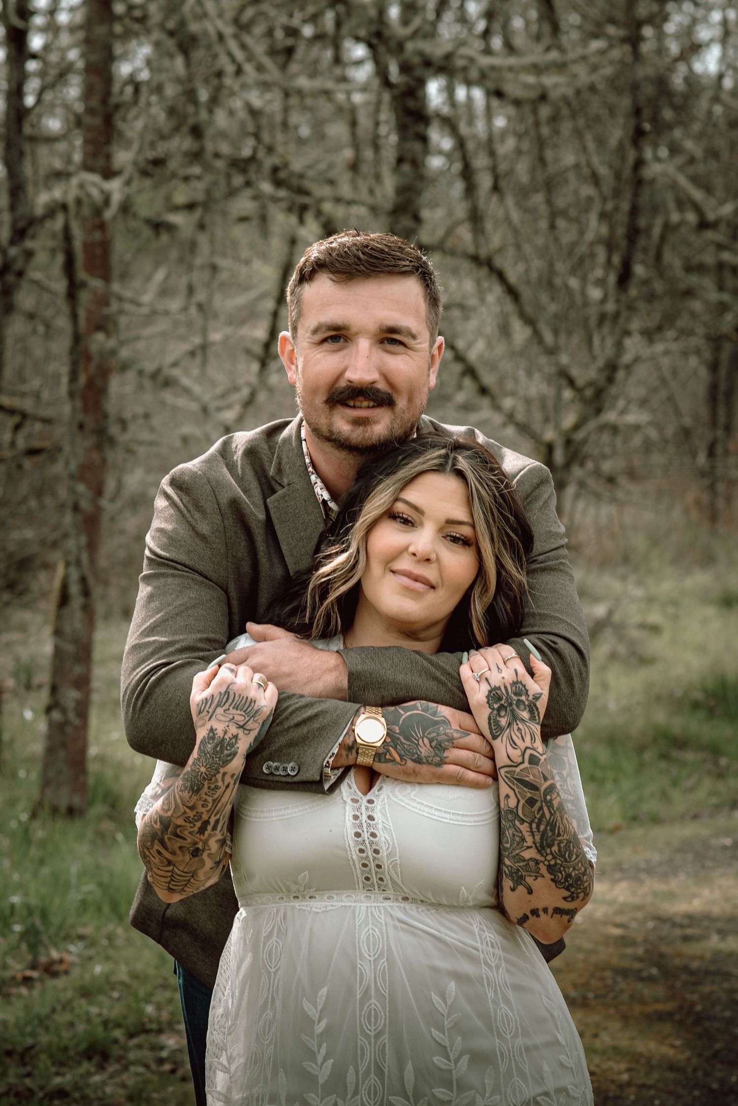 Tattooed couple embracing in a forest, man in olive jacket hugging woman in white dress.