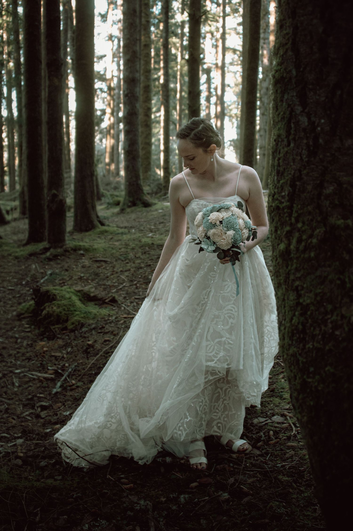 Bride in elegant lace gown holding floral bouquet while standing in a tall pine forest.