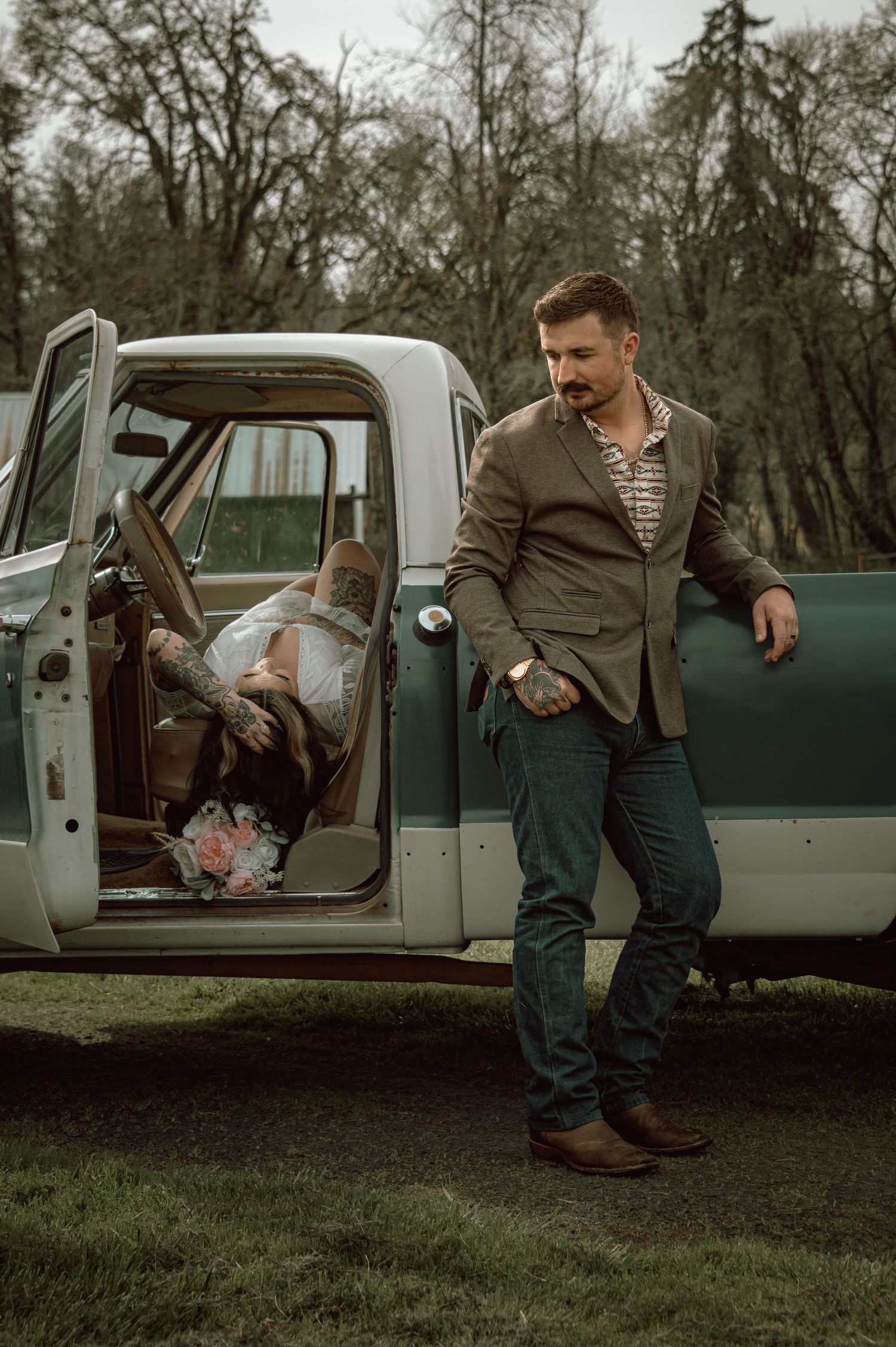 Man leaning against a classic green pickup truck while a woman reclines inside, in a rural outdoor setting.