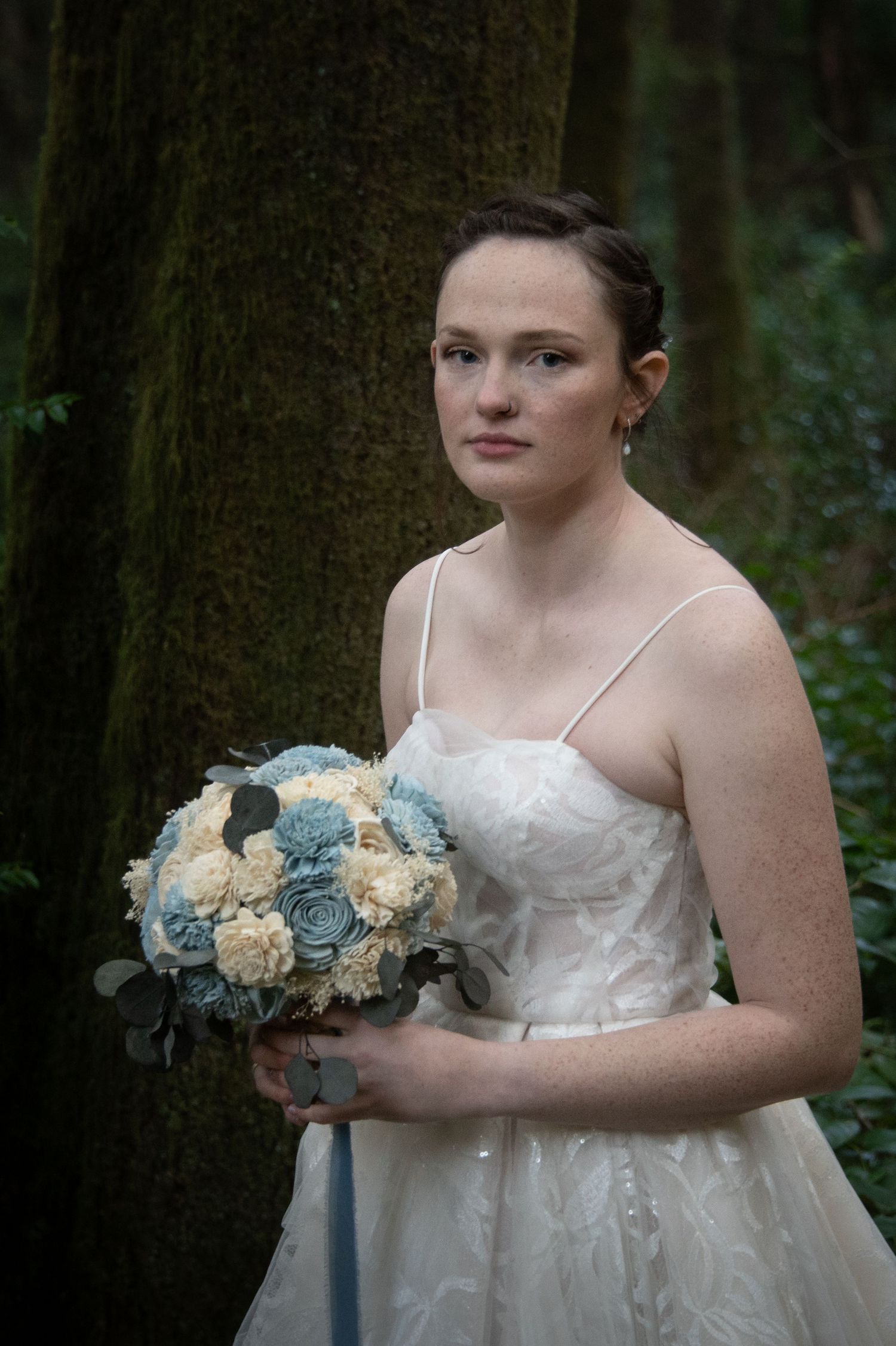 Young bride in white spaghetti-strap gown holding blue and cream bouquet, standing before mossy forest trees.