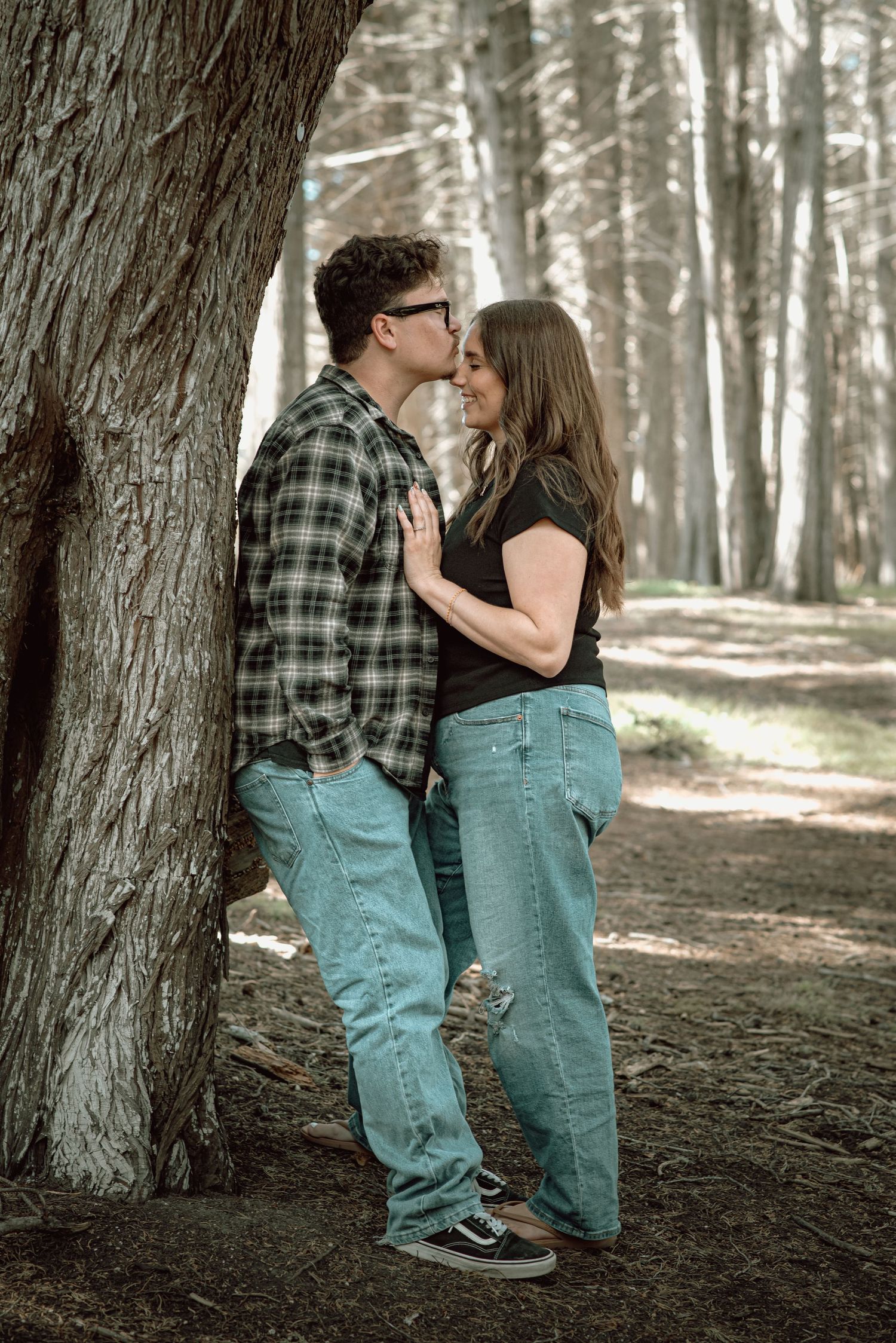 Couple shares a romantic kiss leaning against a tree in a serene forest.