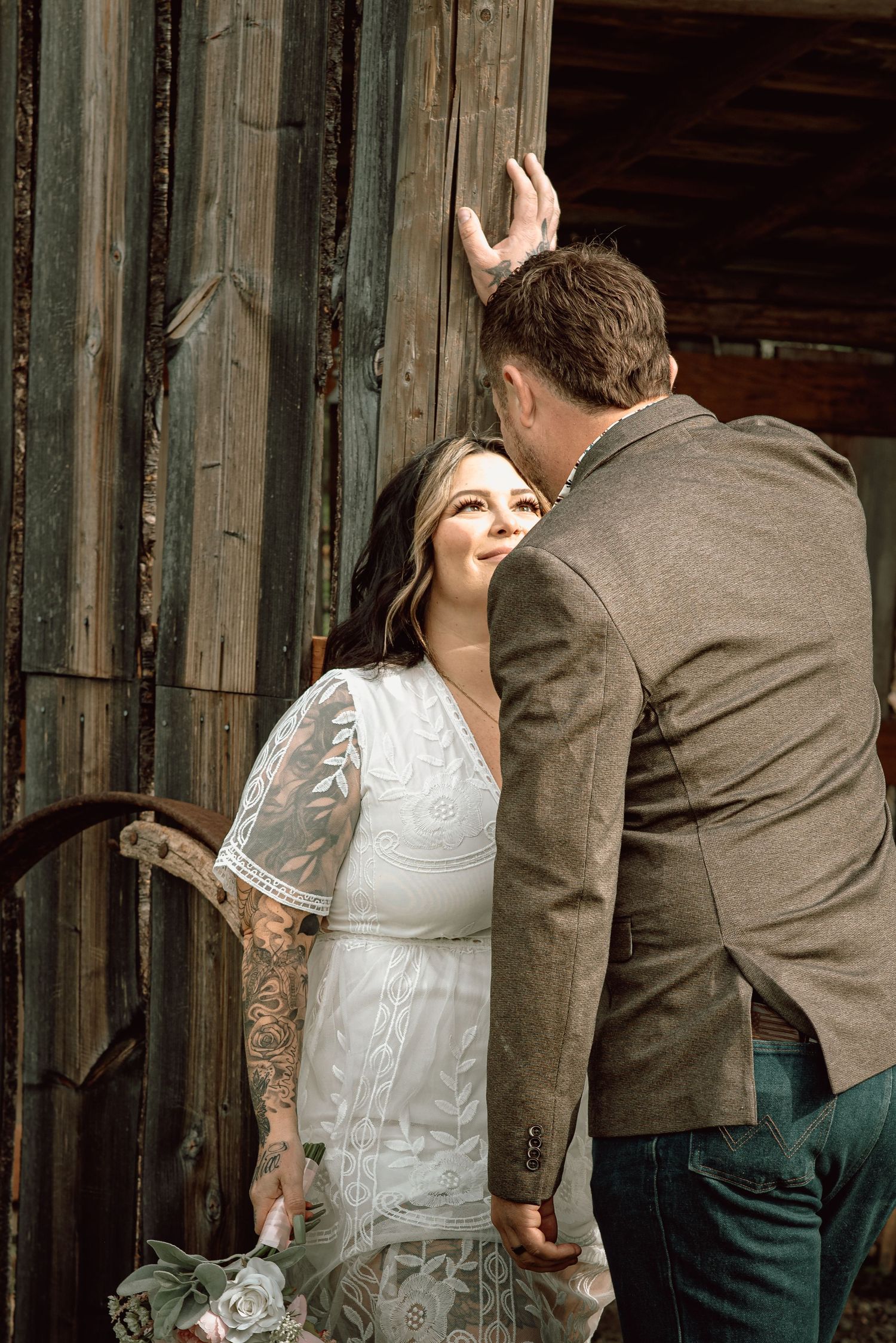 Bride in lace dress with tattoos smiling at groom in olive blazer against rustic barn door.