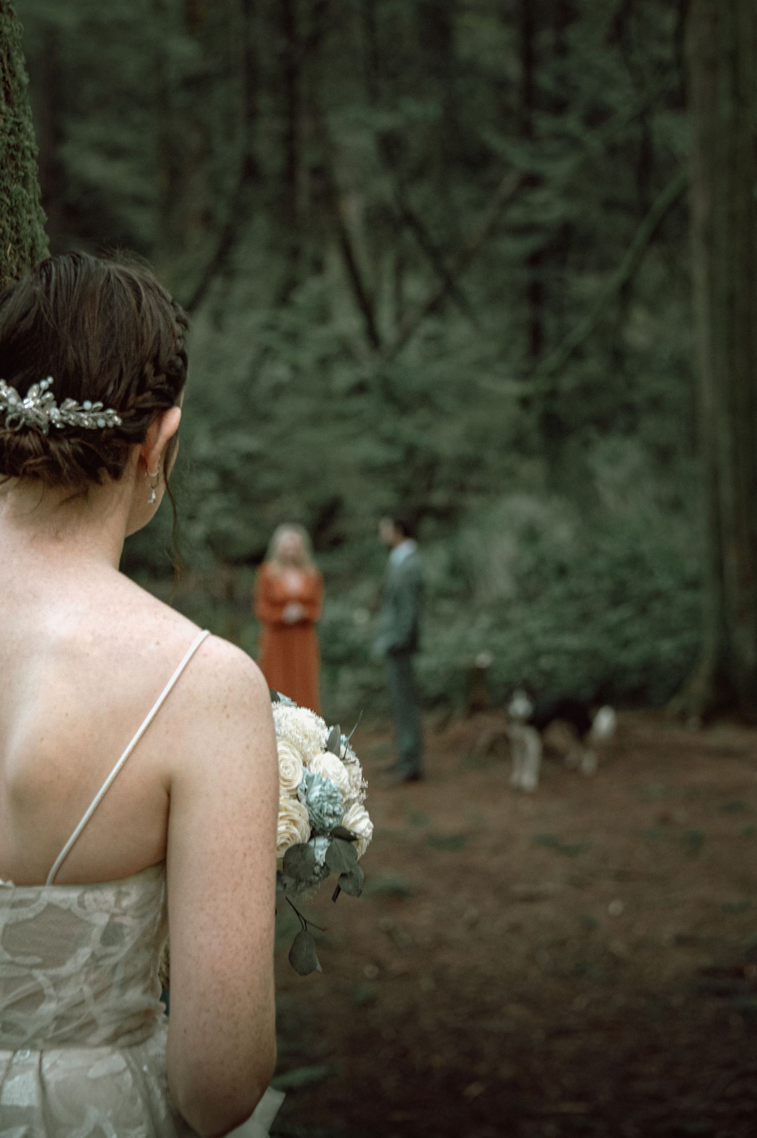 Bride holding blue and white bouquet views officiant and groom standing in a forest clearing ahead.