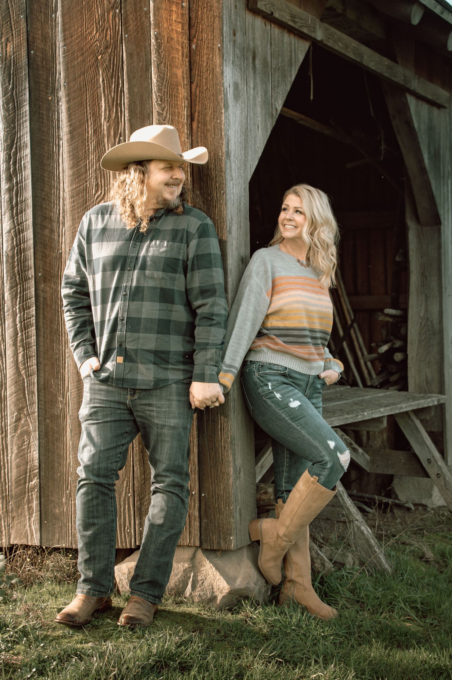 Couple leaning against a rustic barn wall, man in cowboy hat and plaid, woman in striped sweater and boots.